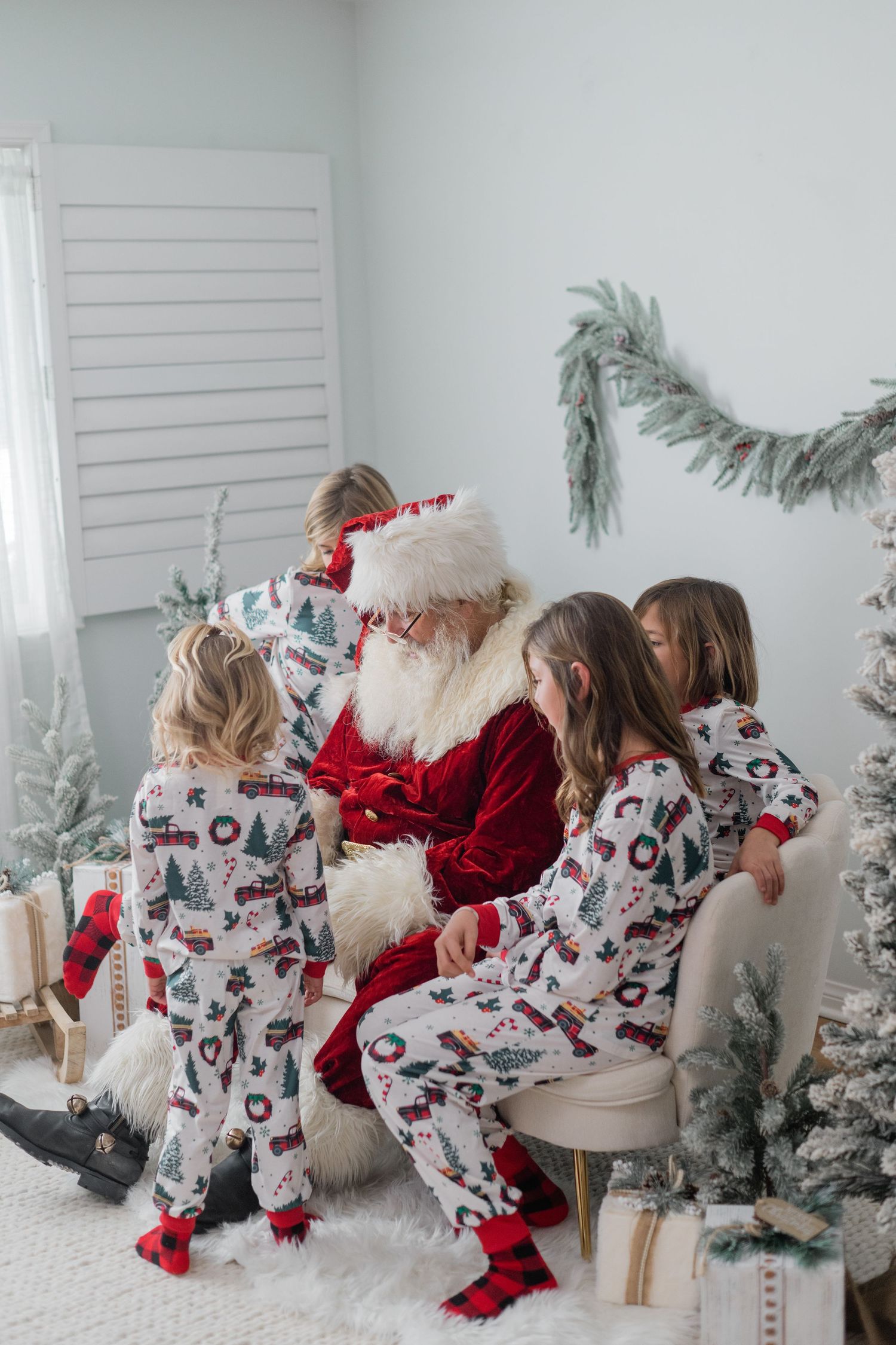 Three children in matching holiday pajamas gather around Santa Claus seated in a chair among Christmas trees.