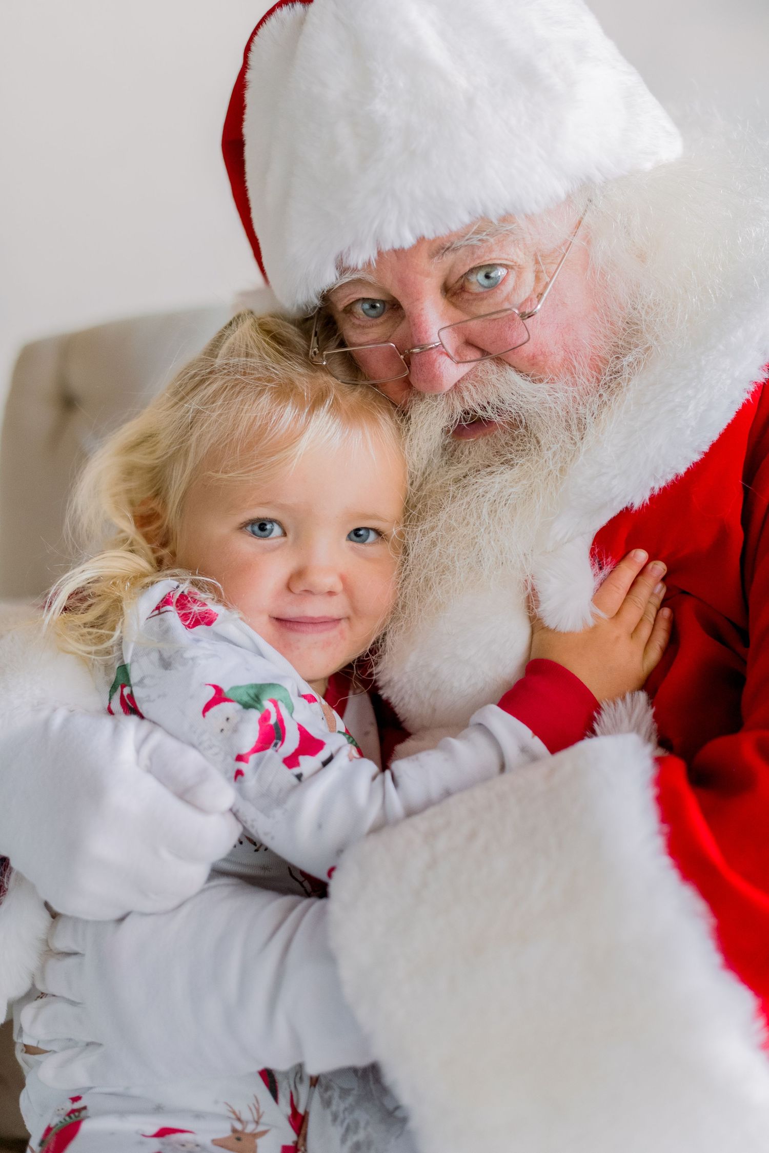 Young blonde girl smiling while sitting on Santa Claus's lap during a festive Christmas portrait session.