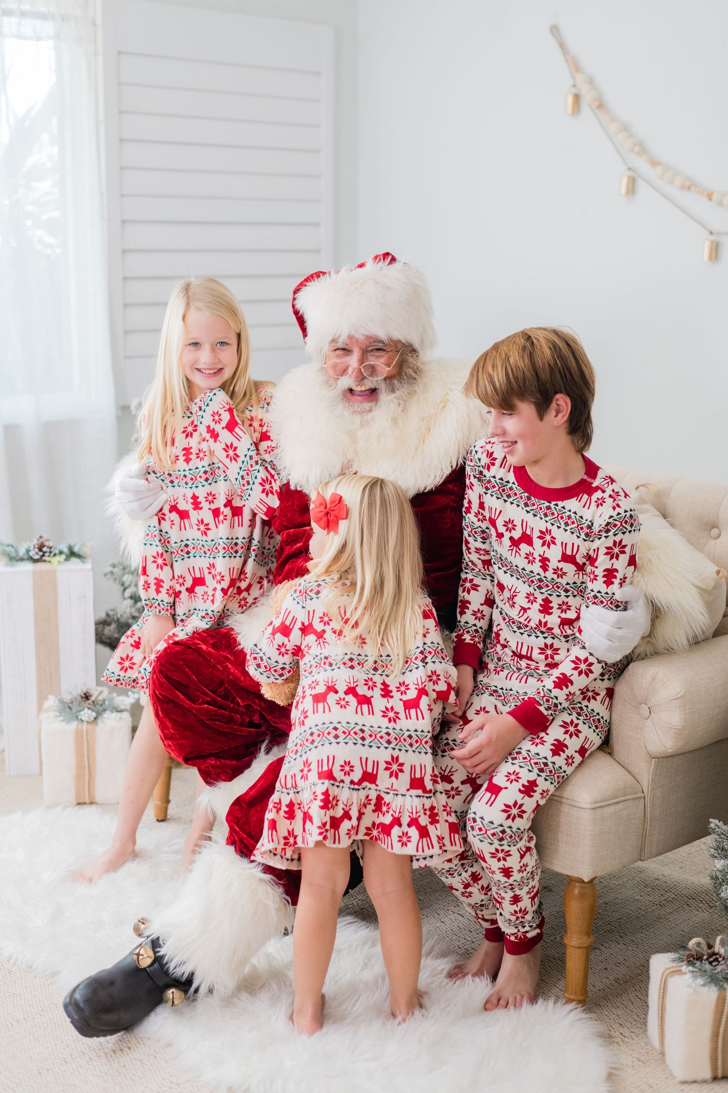 Three children in matching holiday pajamas sit and smile with Santa Claus in a bright festive room.