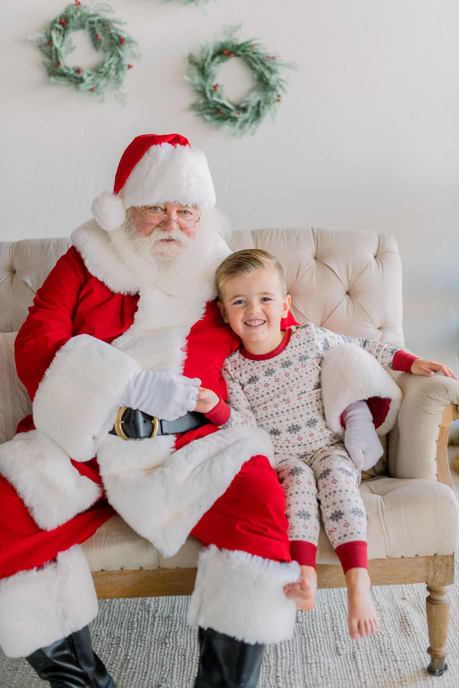 A smiling child in holiday pajamas sits with Santa Claus on a beige tufted sofa with Christmas wreaths on the wall.