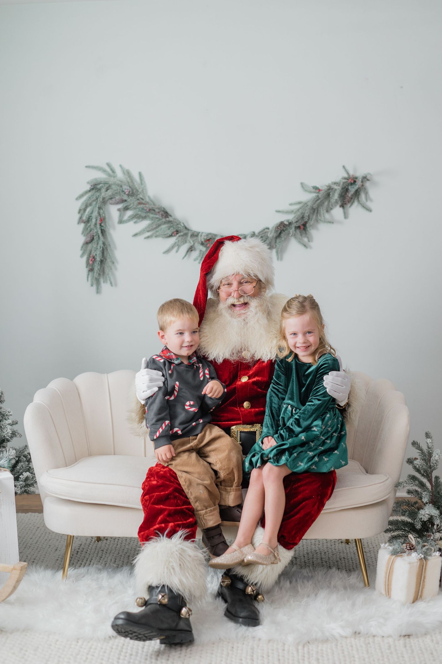 Two children sit with Santa Claus on a cream sofa surrounded by Christmas decor and small trees.