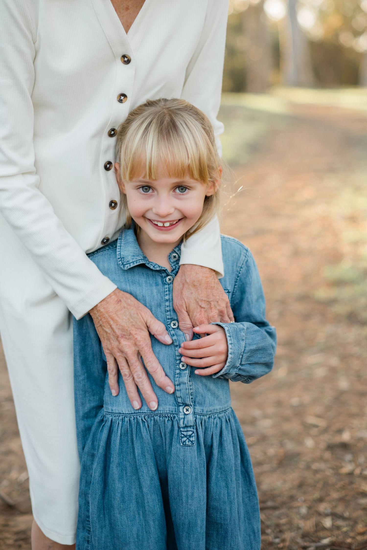 A smiling blonde girl in a denim dress stands outdoors, held gently by an older woman in a white dress.