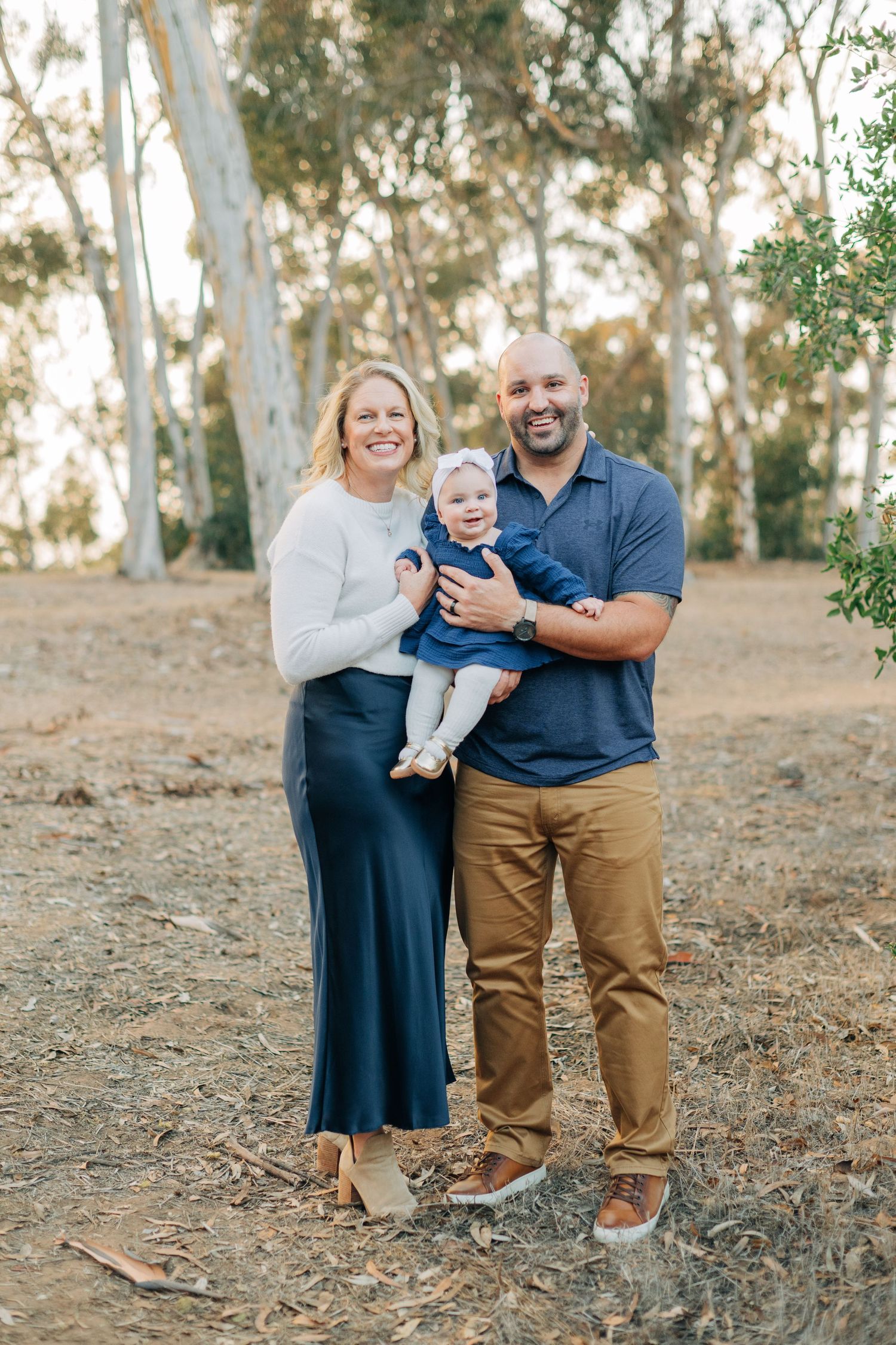 A smiling family of three poses outdoors among eucalyptus trees, parents holding their baby dressed in navy blue.