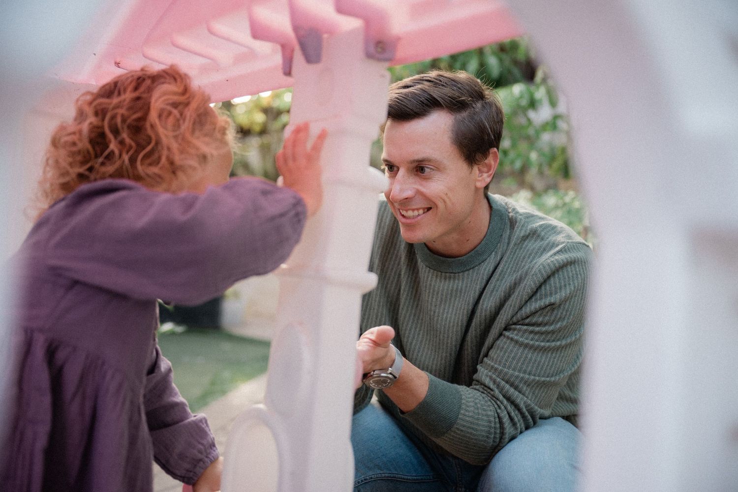 A smiling man in a green sweater kneels to interact with a curly-haired child near a pink playhouse outdoors.