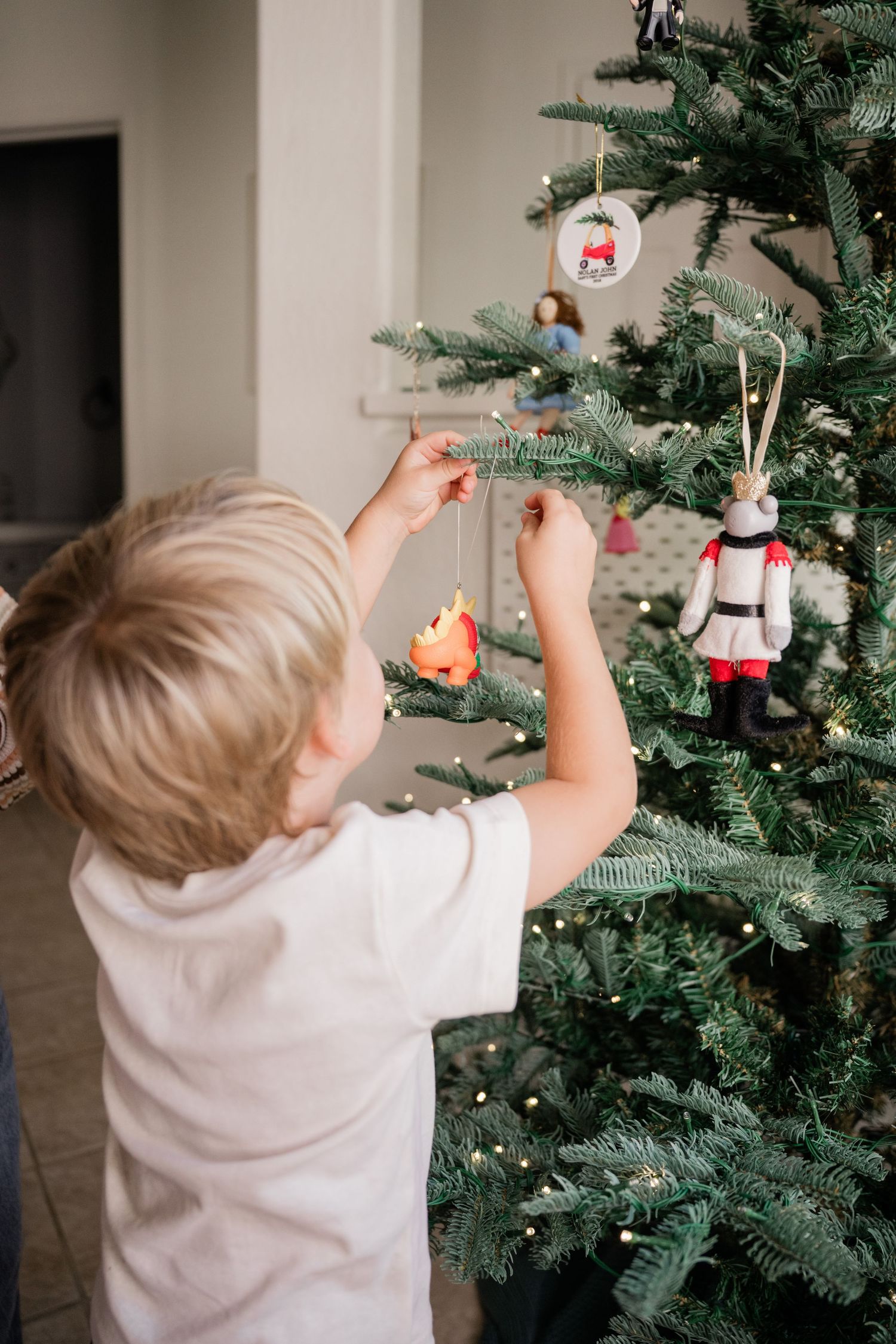 Blonde toddler boy decorating a Christmas tree with colorful ornaments.