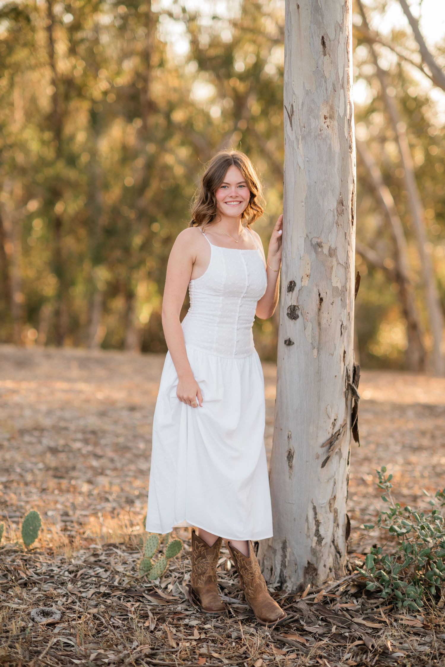 Young woman in white midi dress and cowboy boots smiling beside a tree in a sunlit eucalyptus forest.