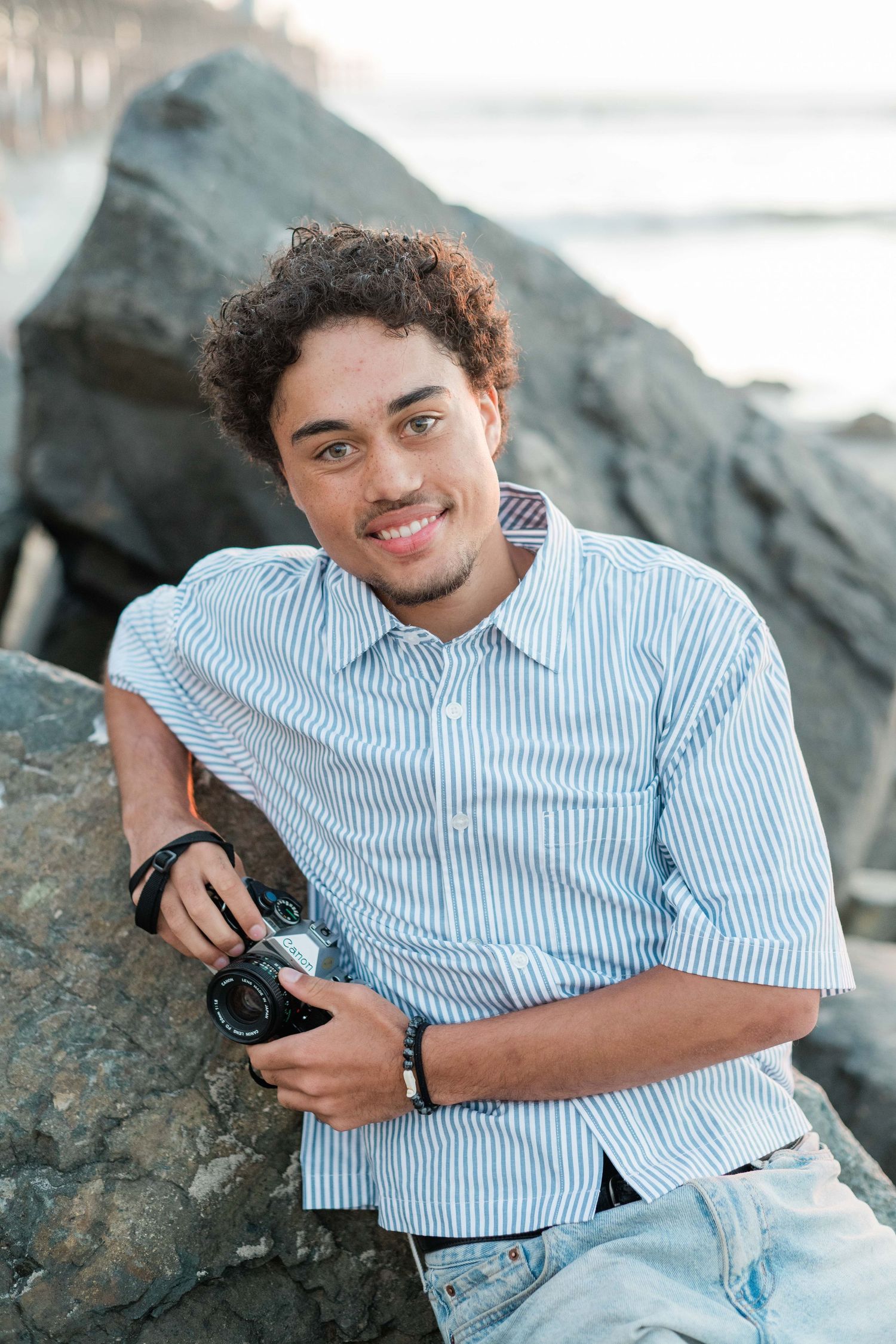 Young man with curly hair holding a vintage camera while leaning against large rocks at the beach, smiling.