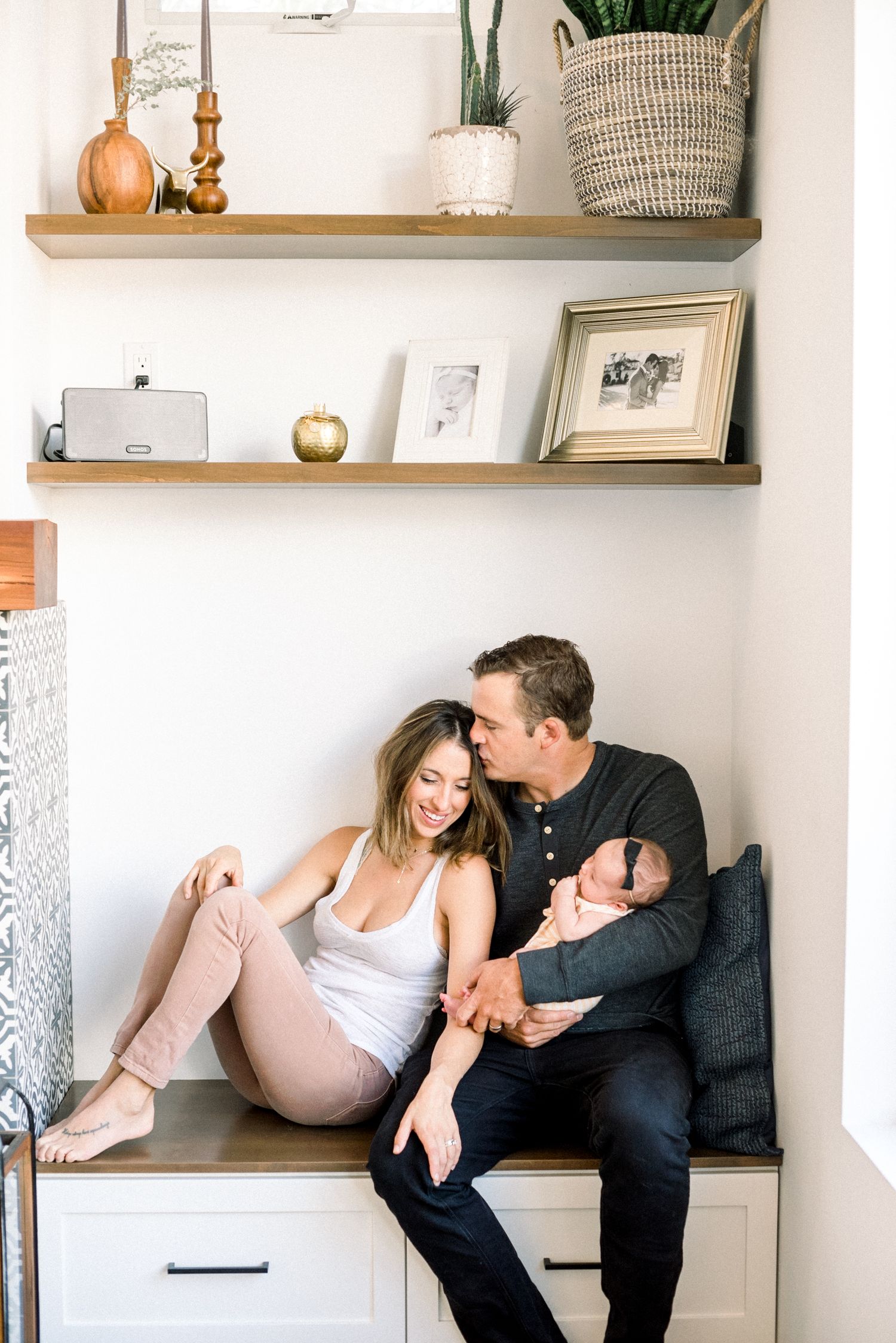 Couple sitting on built-in bench with shelves, sharing a tender moment while holding their newborn baby.