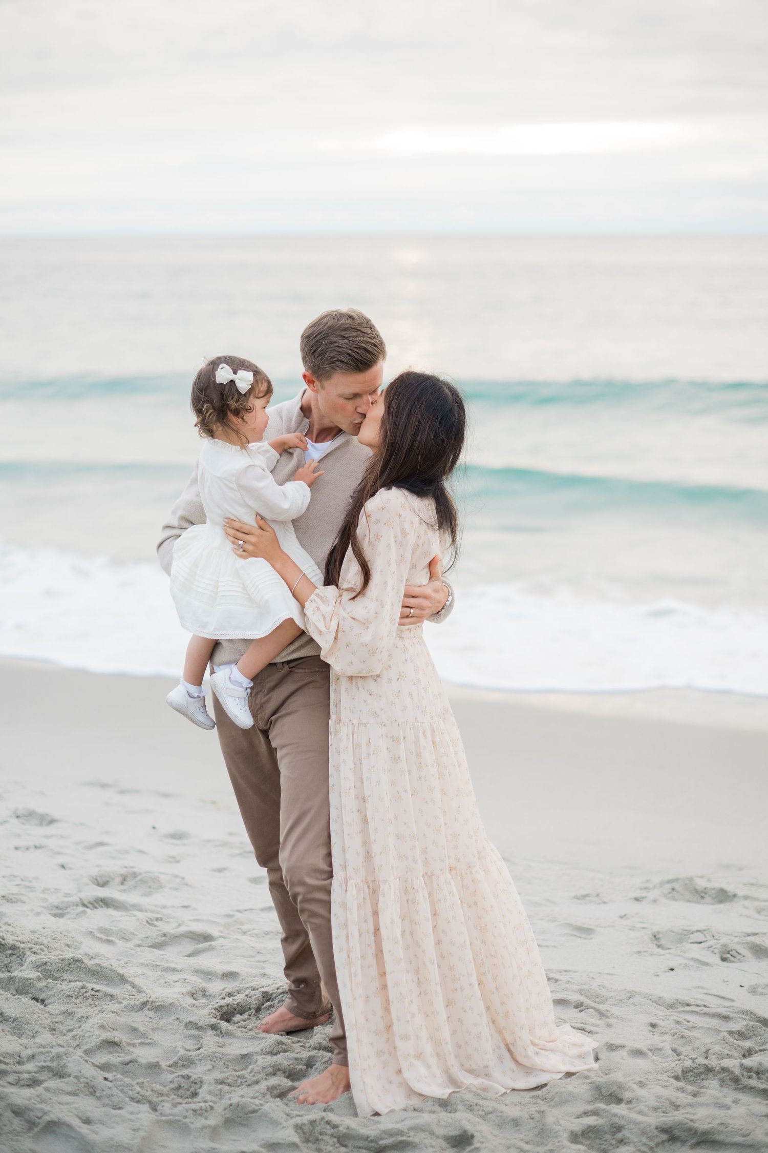 A couple shares a kiss on a sandy beach while holding their baby girl, dressed in elegant neutral outfits.