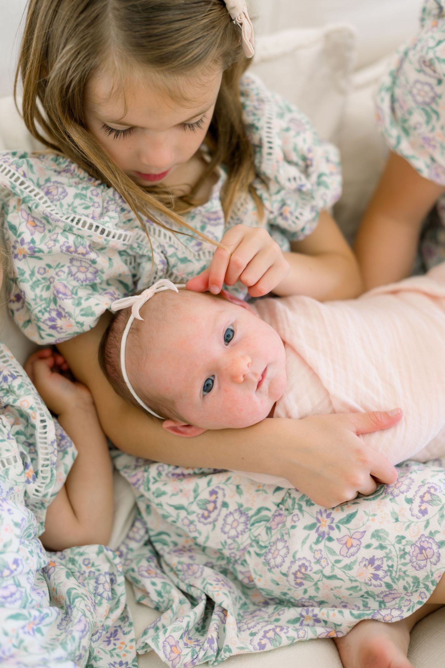 Young girl in floral dress tenderly holds newborn baby wrapped in pink swaddle with headband.