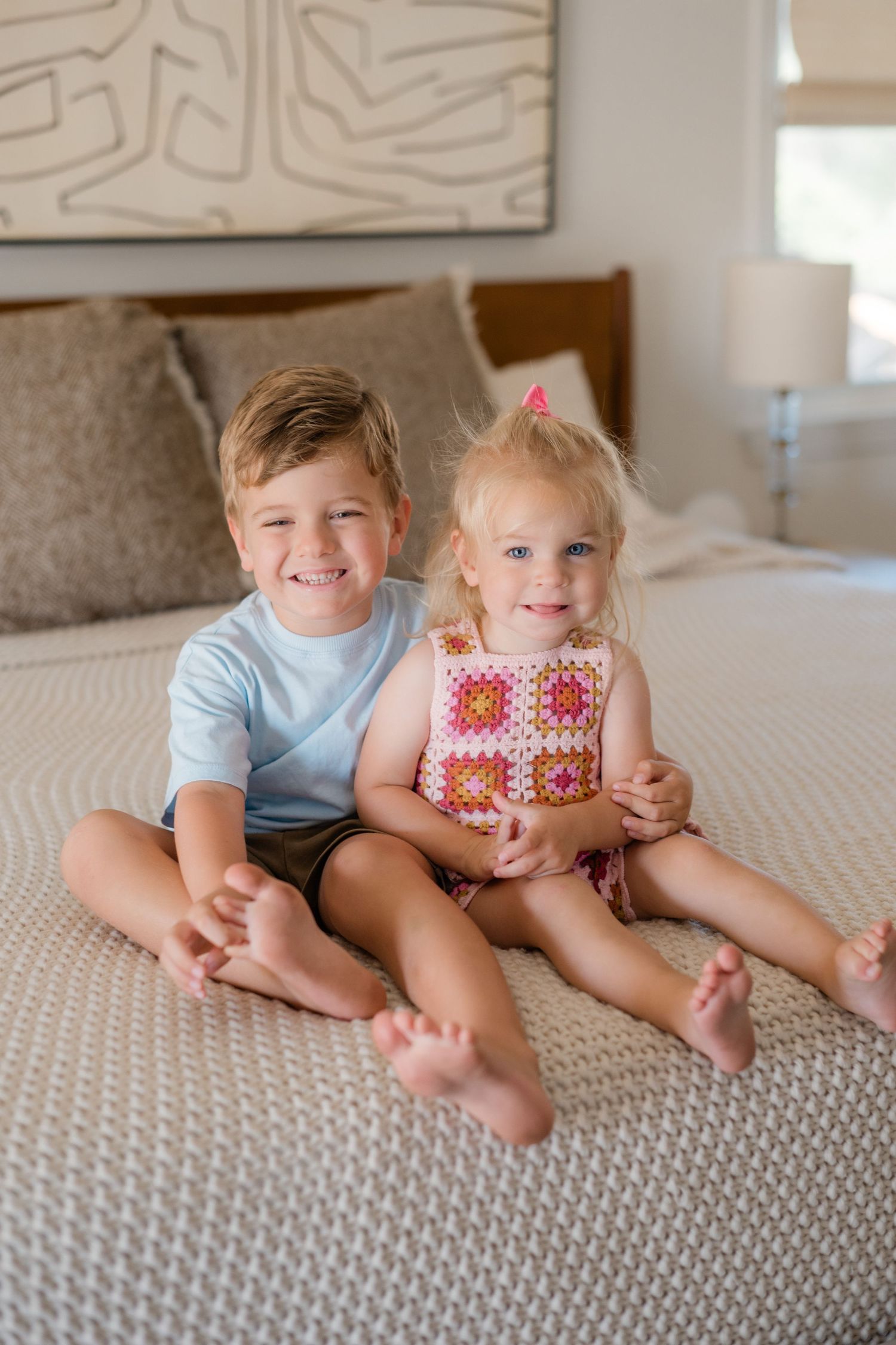 Two young siblings, a boy and girl, sitting together smiling on a bed in a bright bedroom.