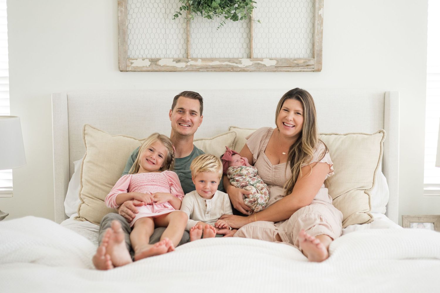 Happy family of five posing on a white bed, parents smiling with three young children including a newborn baby.