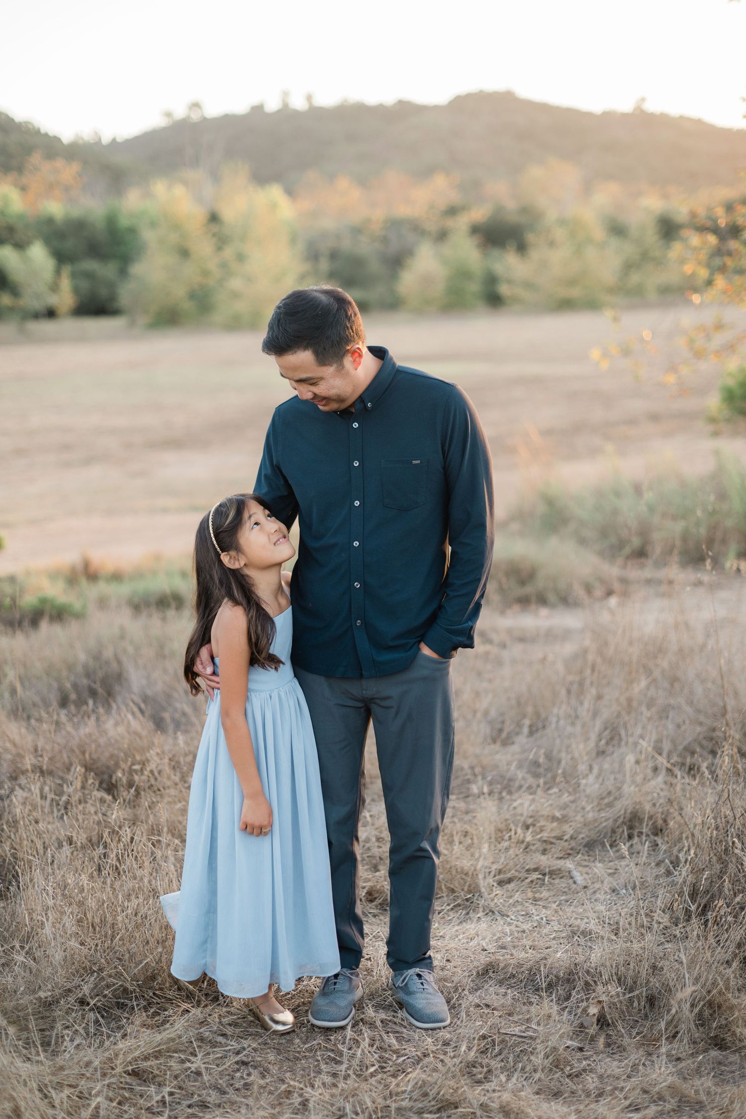A father and young daughter share a tender moment in a golden field, she wears a light blue dress, he a navy shirt.