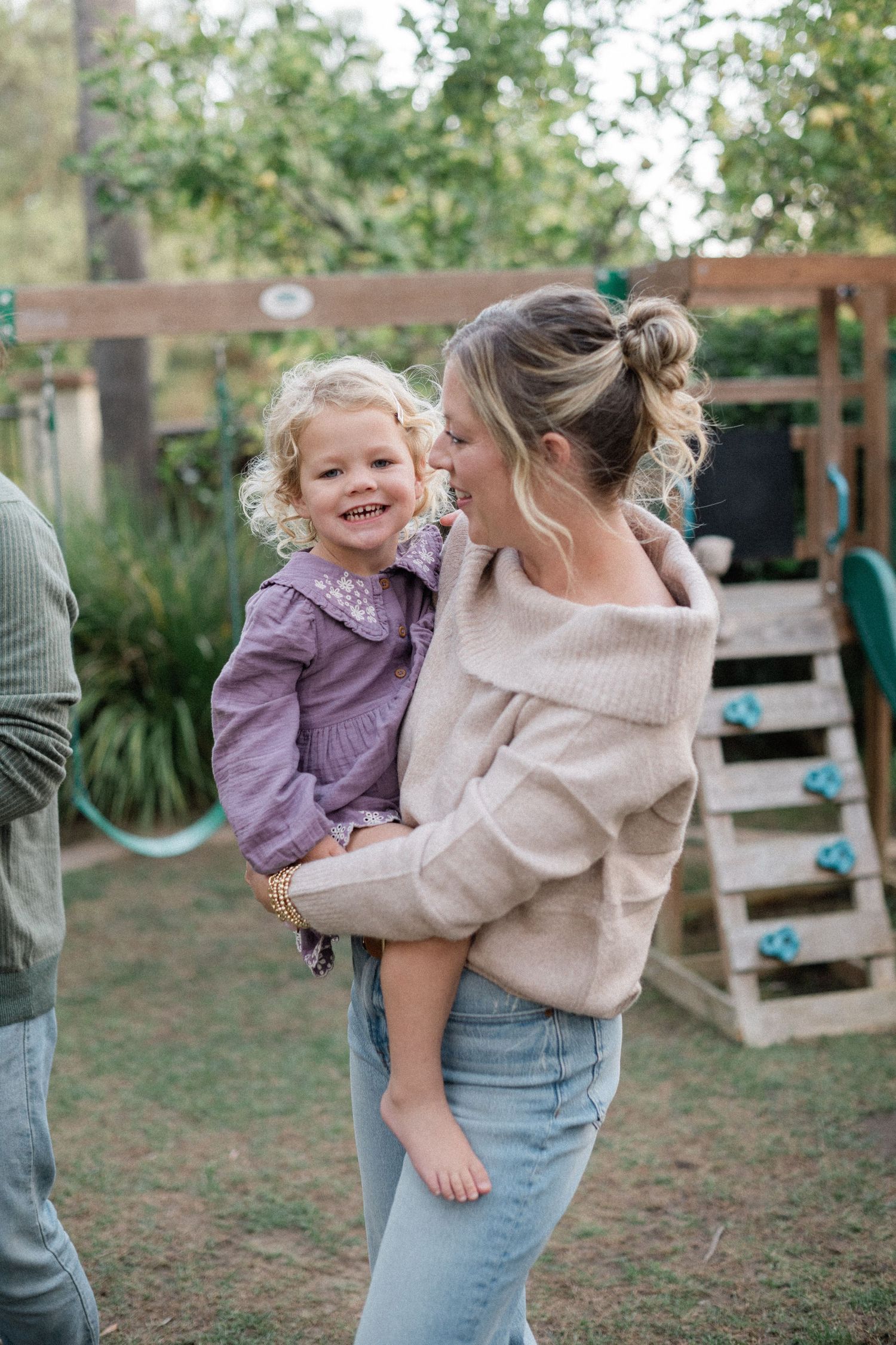 Happy toddler girl in purple dress being carried and kissed by her smiling mother in a backyard.