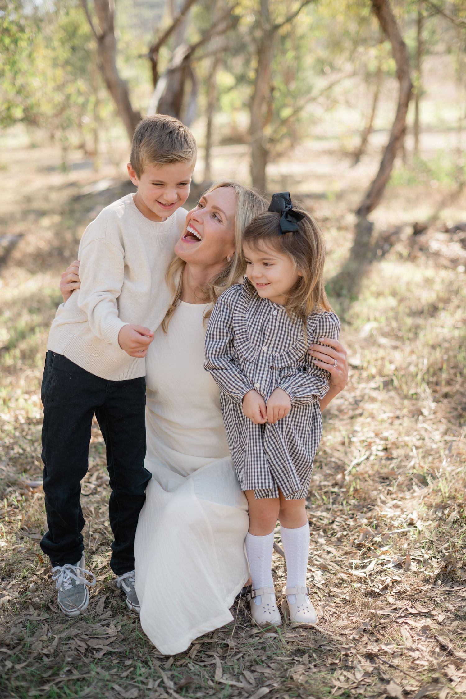 A smiling blonde woman kneels outdoors hugging two young children, a boy in cream sweater and girl in plaid dress.
