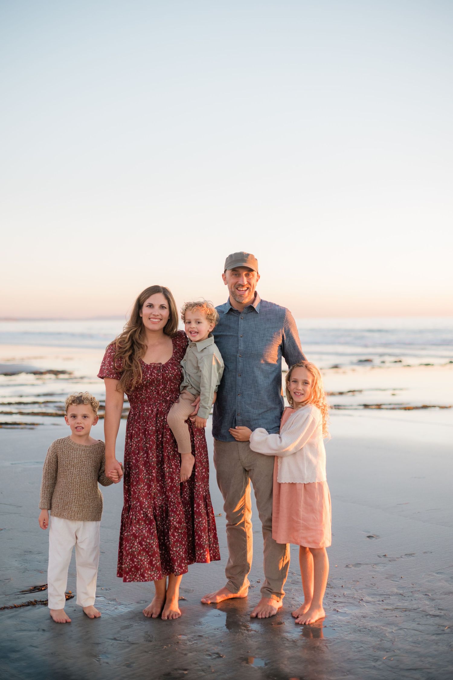 Family of five posing barefoot on a sandy beach at sunset, with ocean waves in the background.