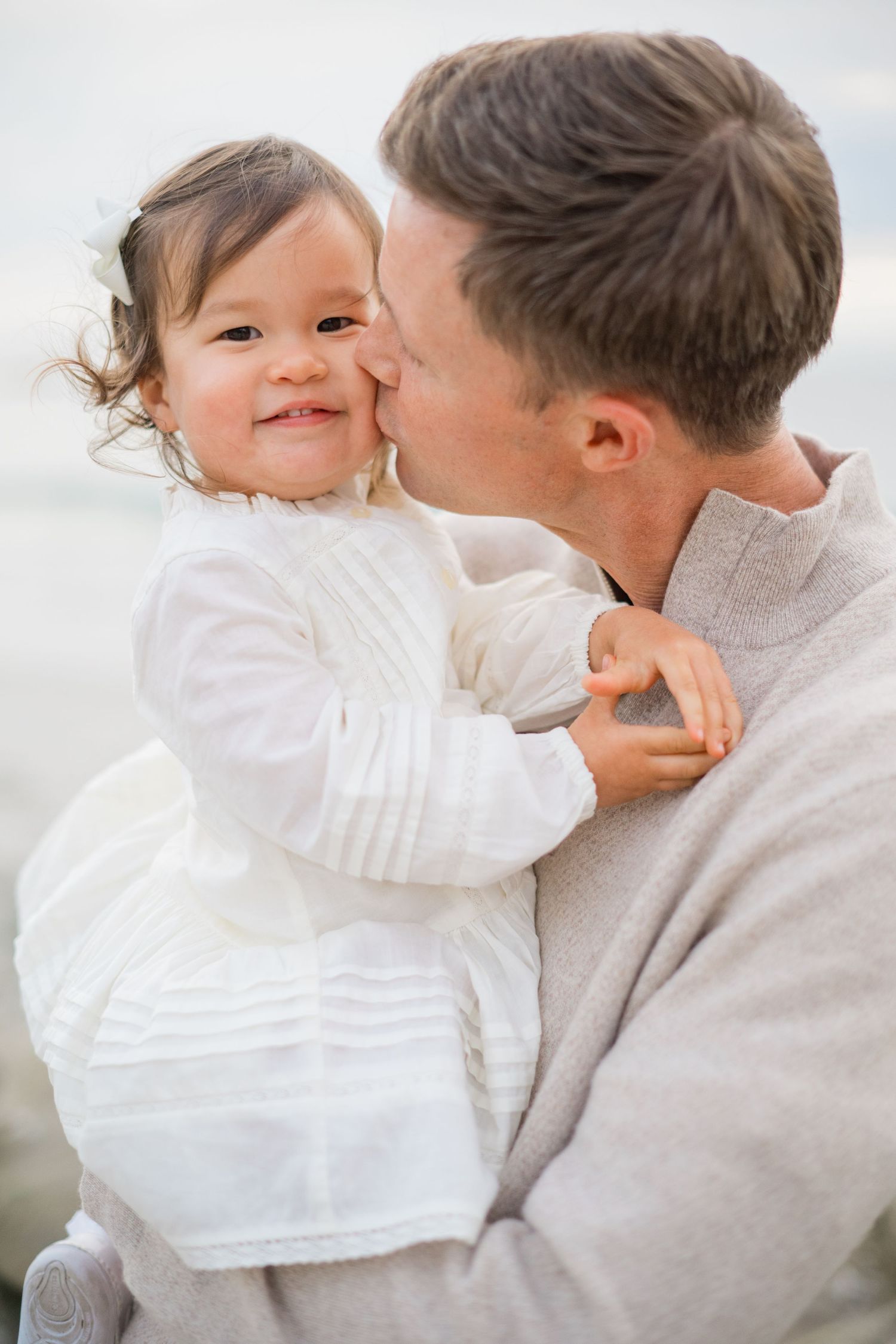 A father tenderly kisses his smiling toddler daughter in a white dress on a beach.