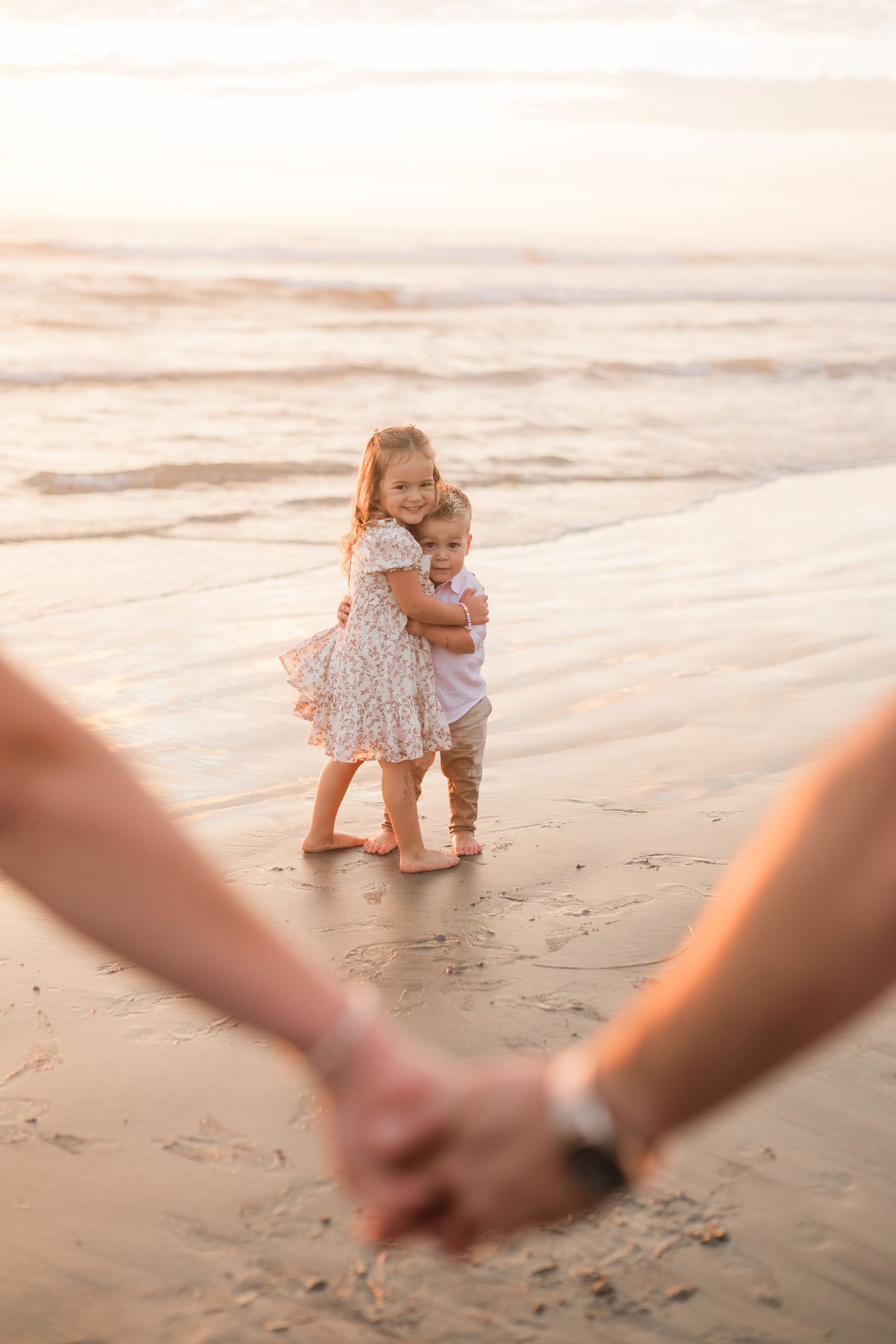 Two young children hug on a sunny beach at sunset while parents hold hands in the foreground.