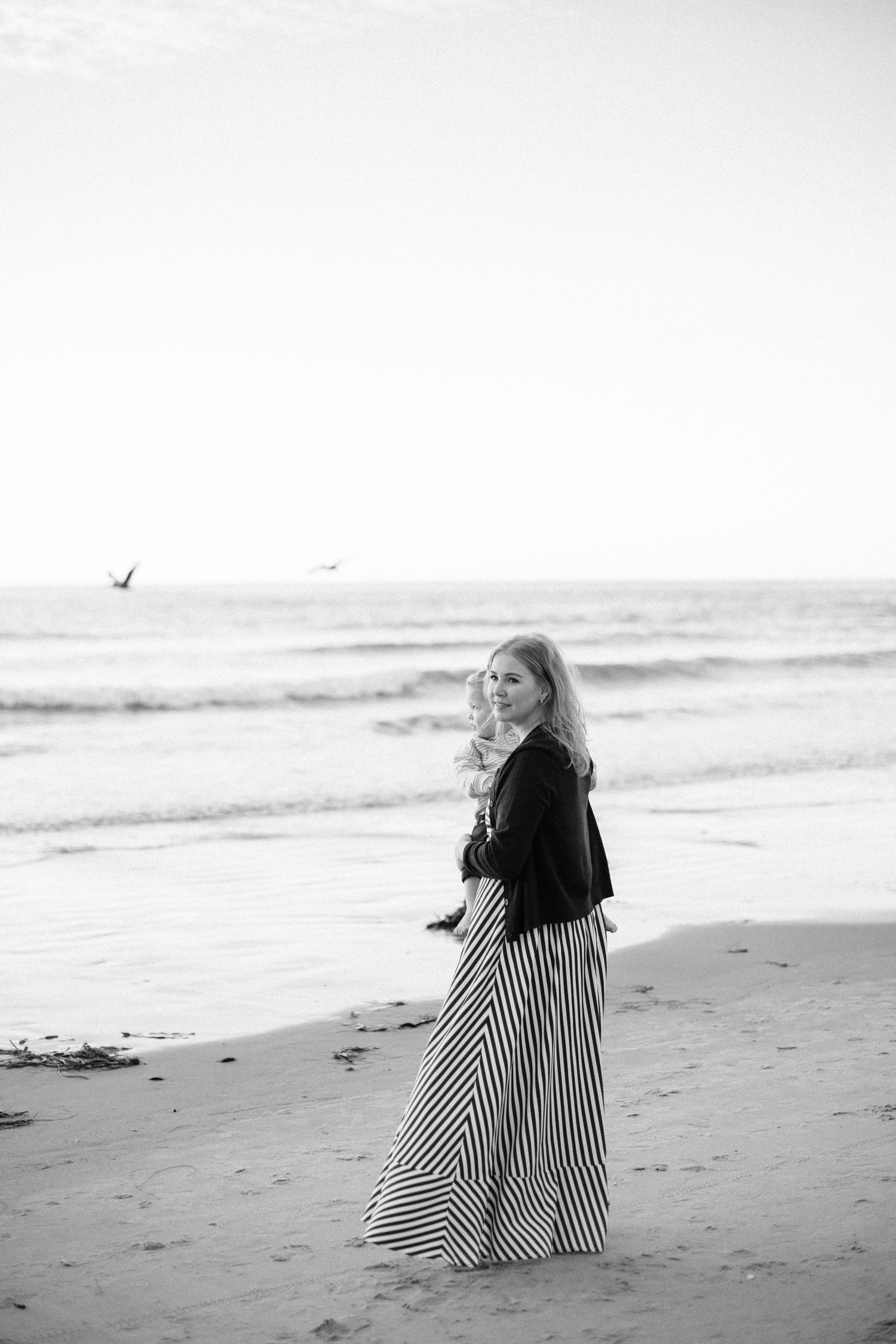 Woman in striped maxi skirt standing on sandy beach near ocean waves in black and white photo.