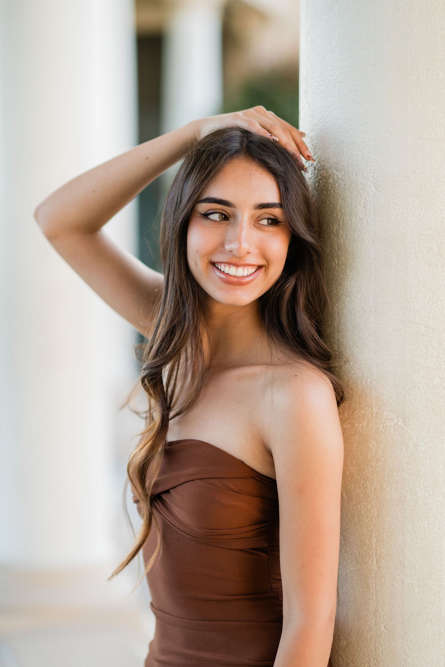 Young woman in brown strapless dress smiling, leaning against a column with hand in hair.