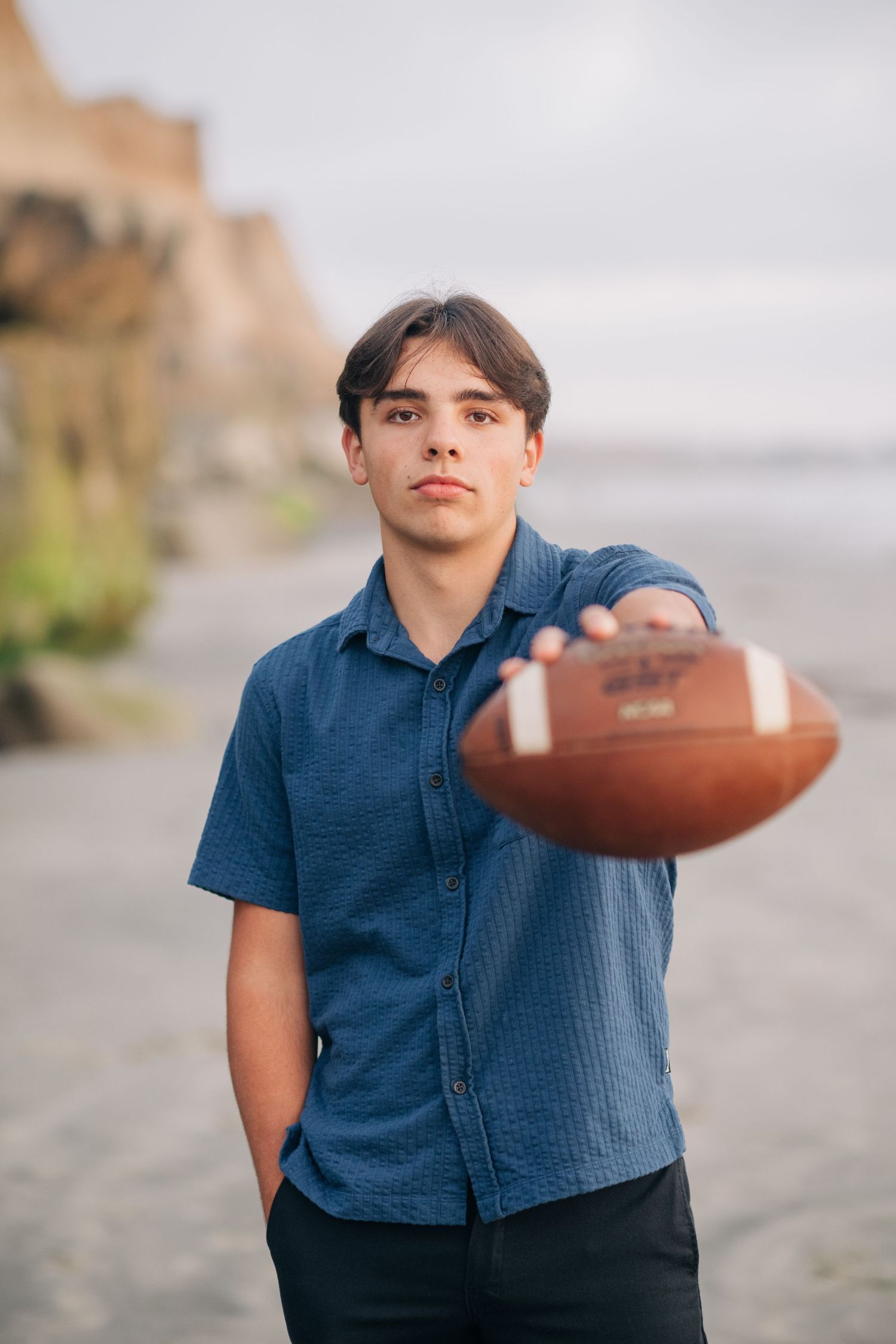 Teen boy in blue shirt extending a football toward camera on a beach with cliffs in background.