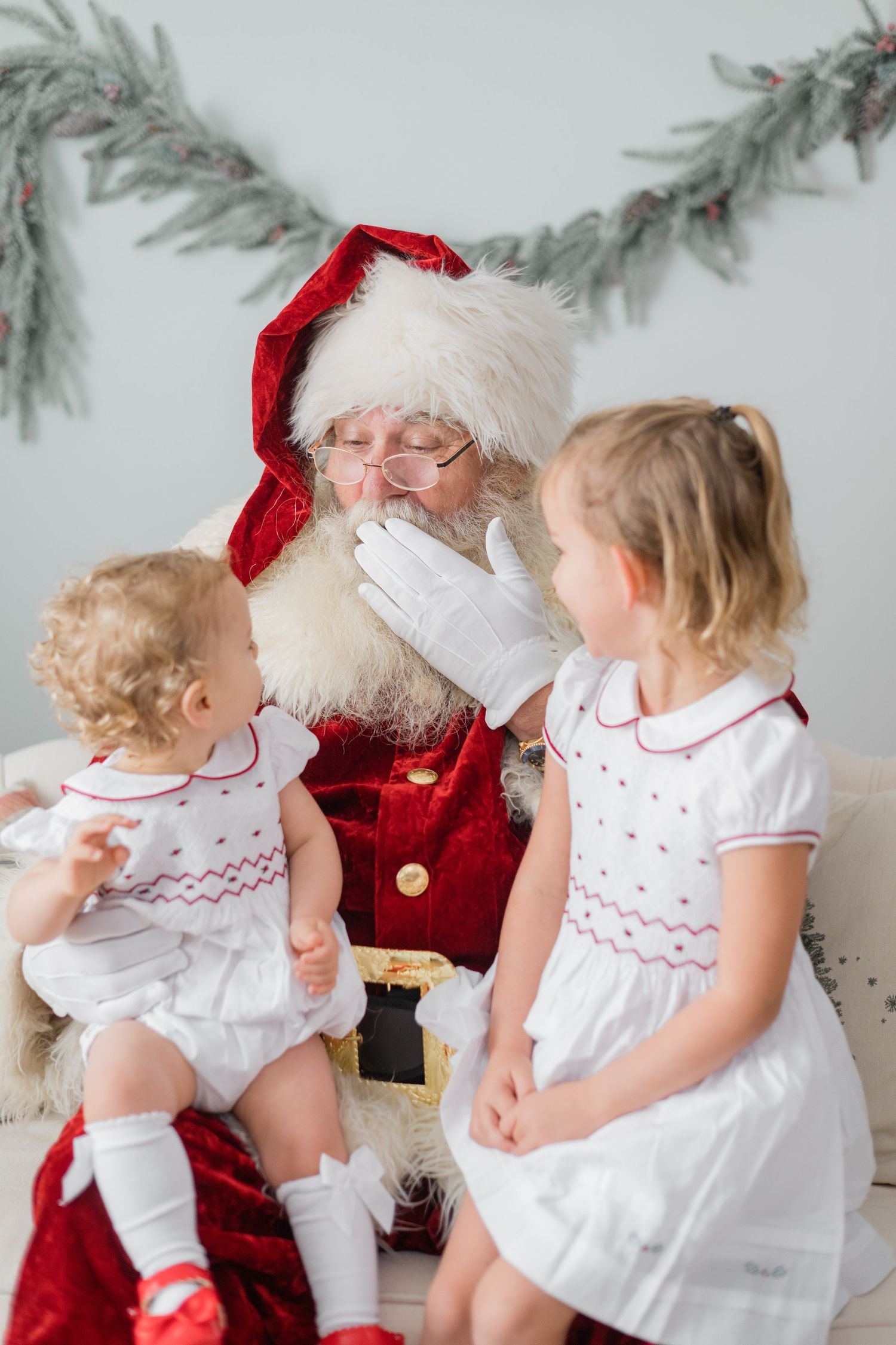 Santa Claus interacts with two young girls in matching white smocked dresses during a festive Christmas portrait session.