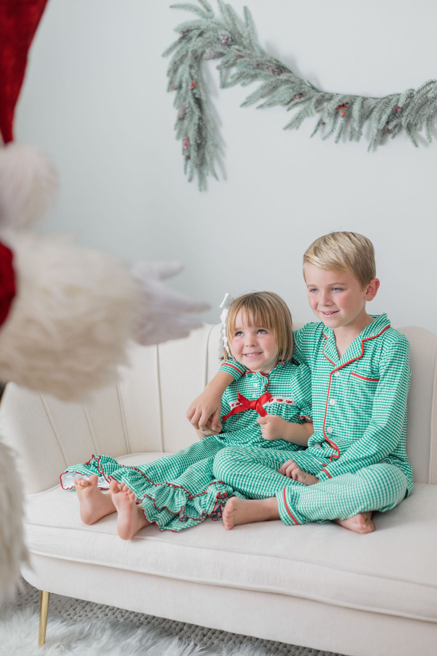 Two siblings in matching green plaid Christmas pajamas sit together on a cream sofa with Santa Claus nearby.