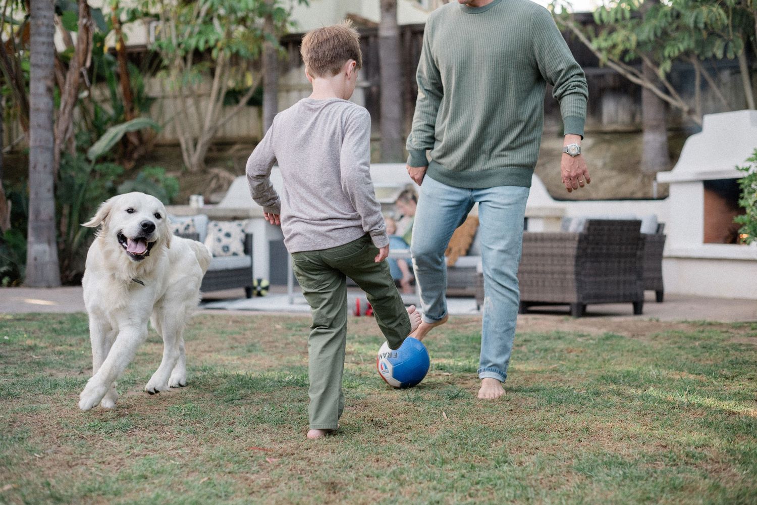 A father and child play soccer with a blue ball in a backyard while a happy golden retriever dog joins the fun.