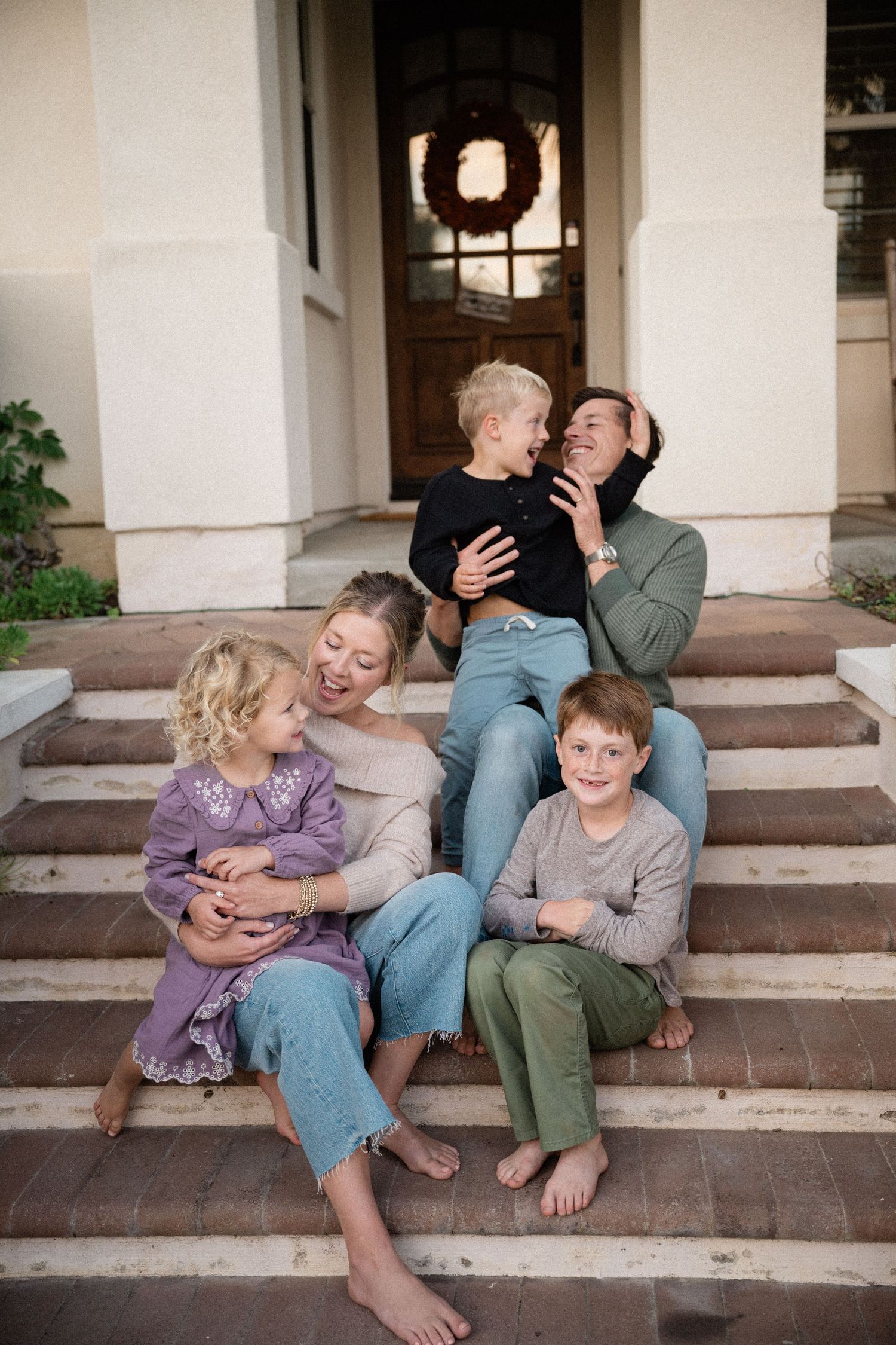 Happy family of five laughing together on front porch steps of a home, barefoot in casual clothing.