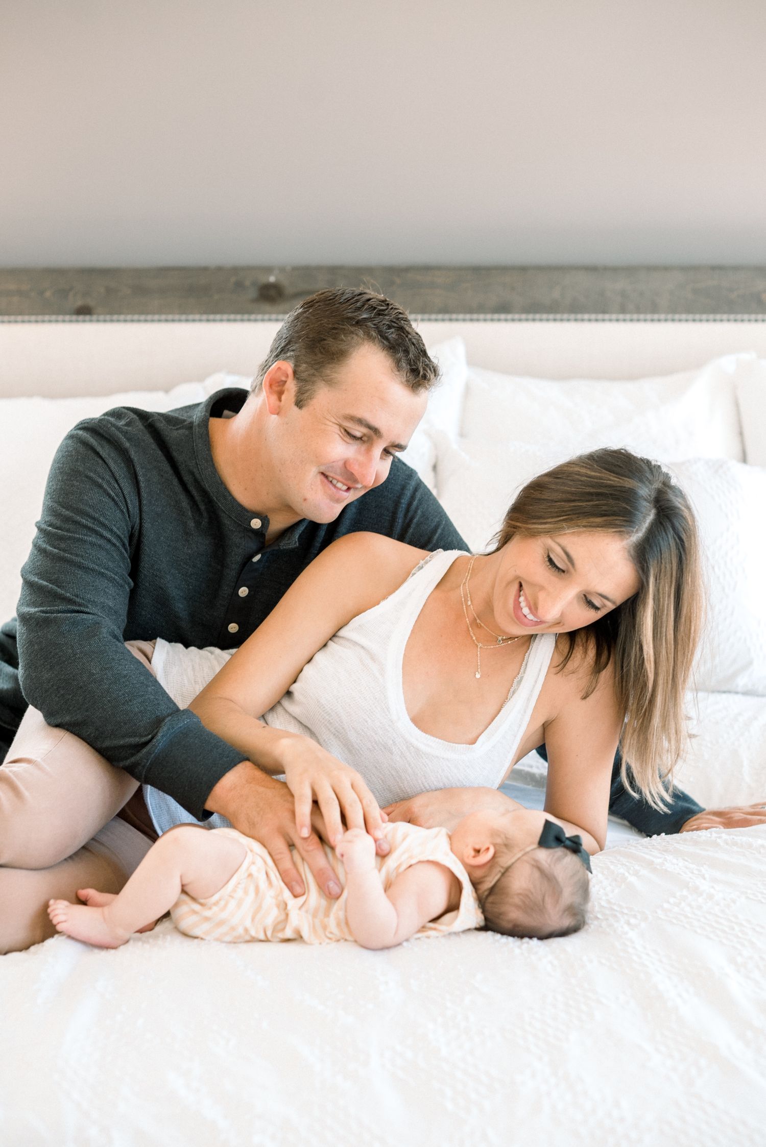 Smiling parents sit on a bed together admiring their newborn baby lying between them.