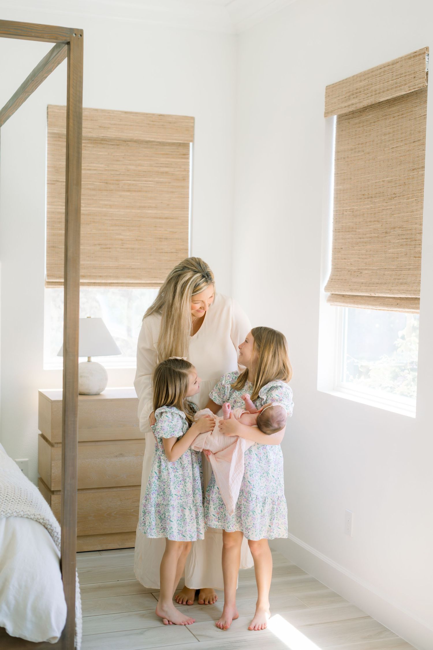 Mother in white dress shows newborn baby to two young daughters in floral dresses in bright bedroom.