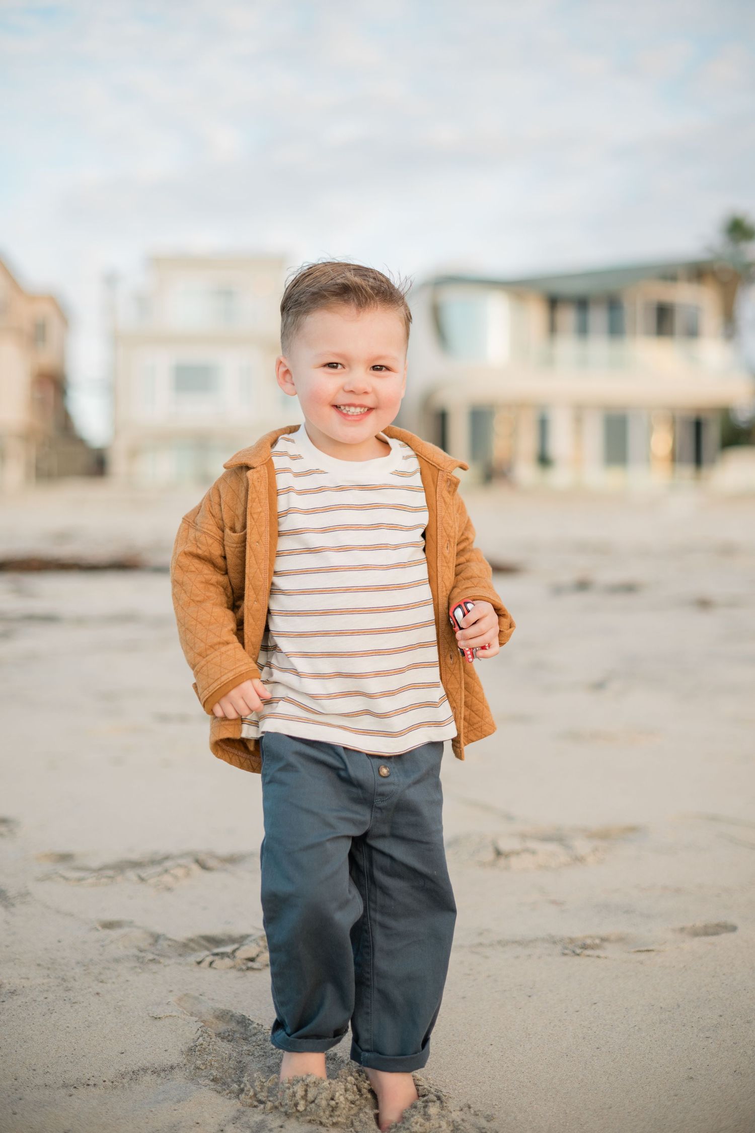 Smiling toddler boy in striped shirt and brown jacket playing on sandy beach with houses in background.