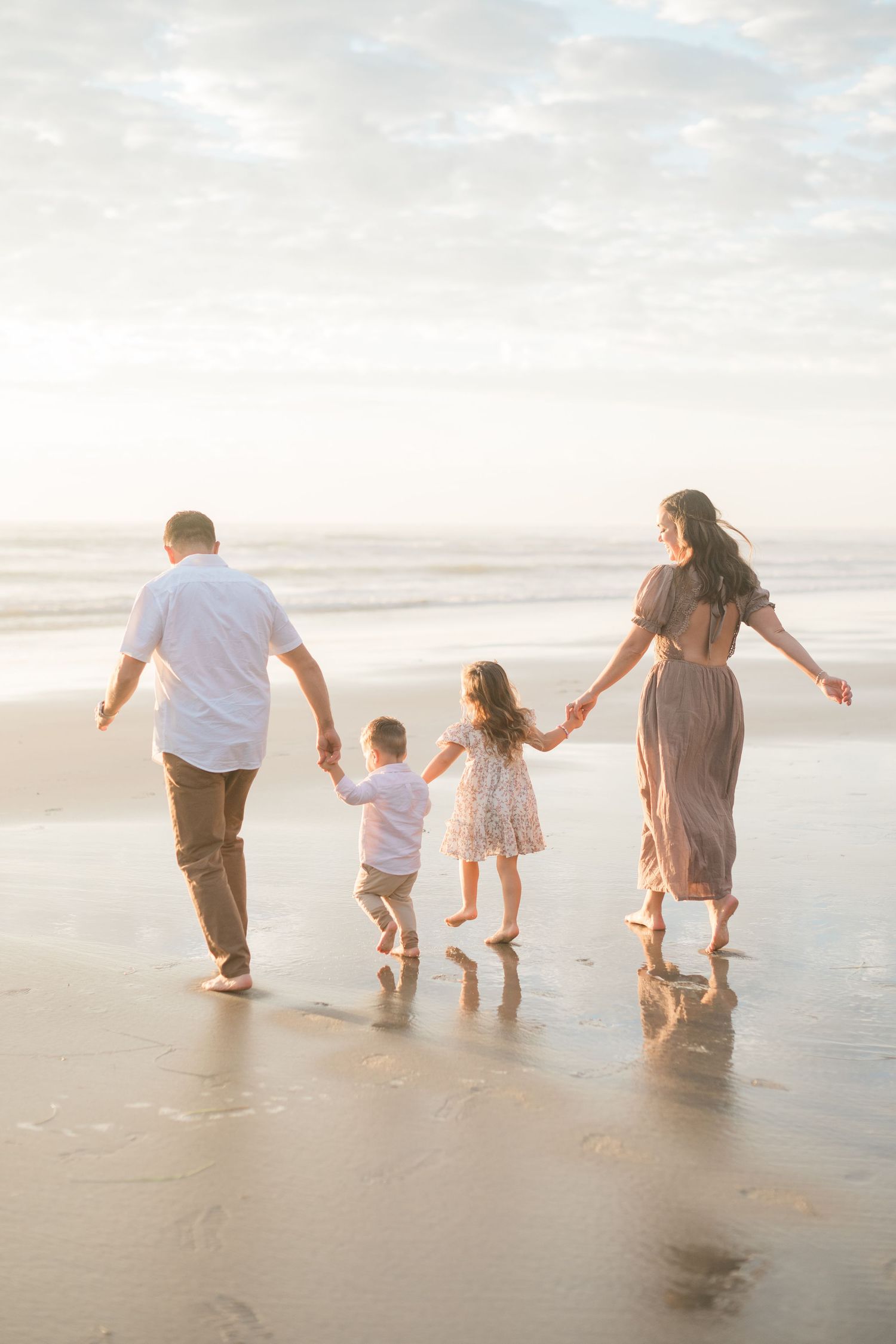 Family of four walking hand-in-hand on a sandy beach at sunset, with two young children between their parents.