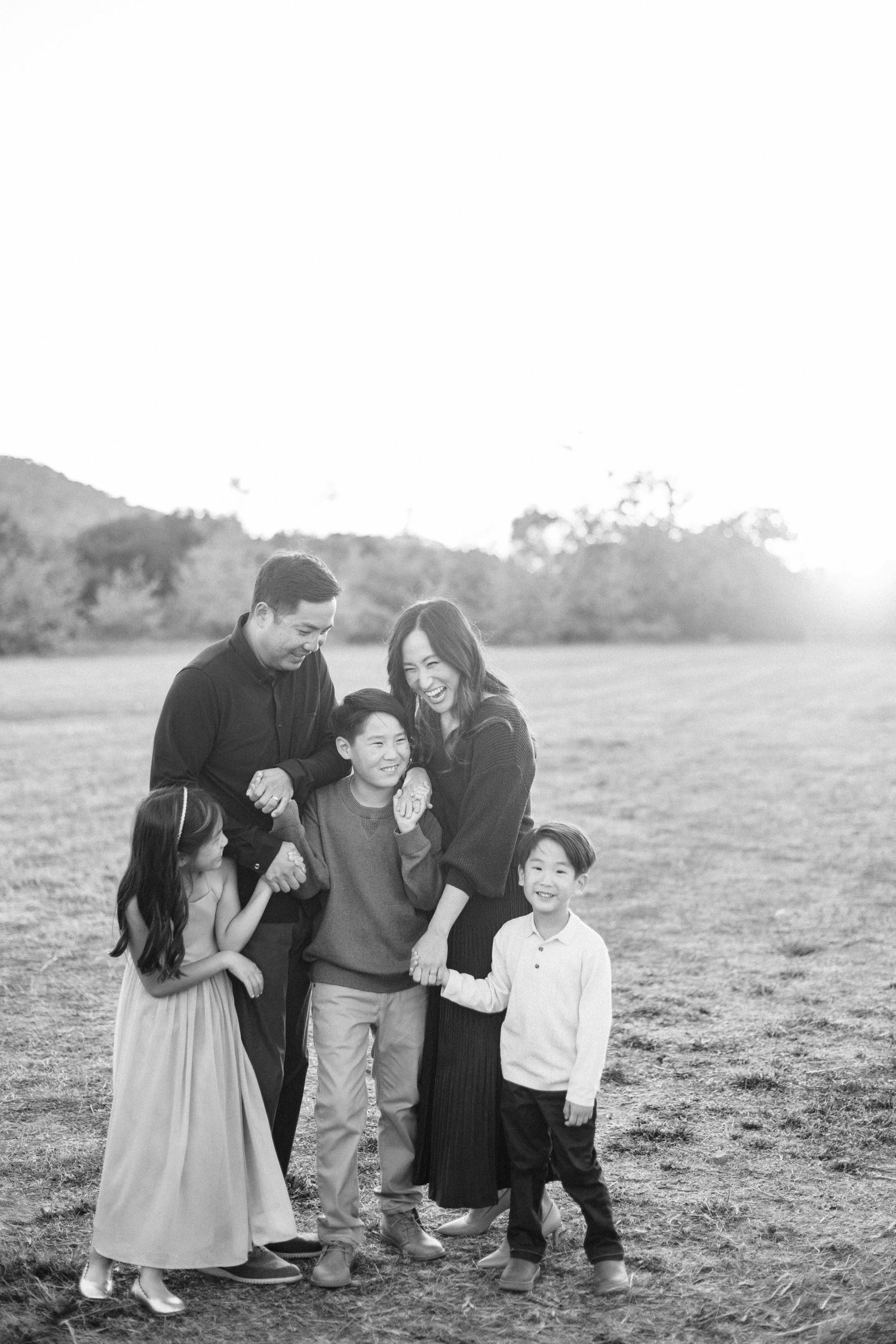 A black and white portrait of a happy Asian family of five embracing in an open field at golden hour.