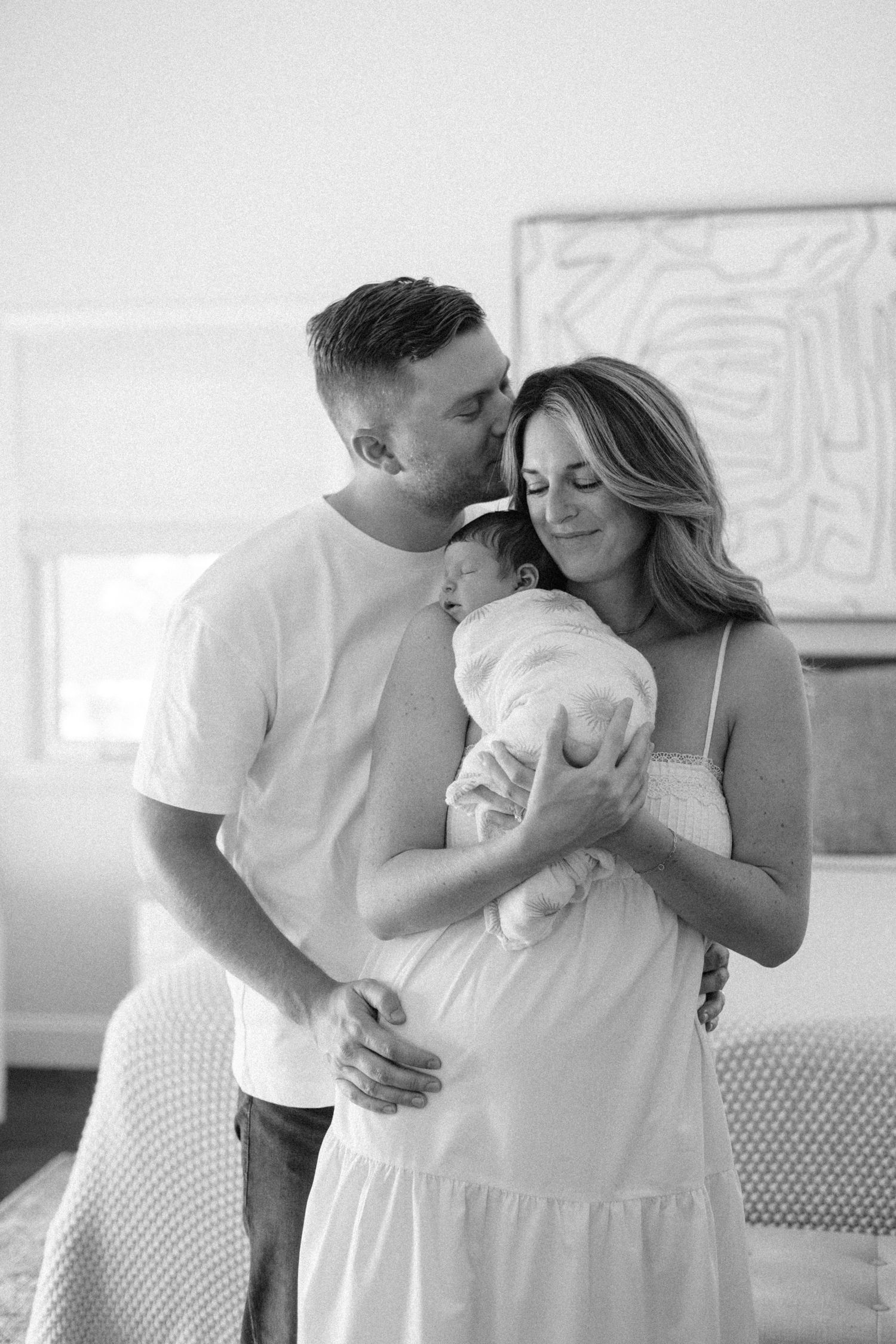 Black and white photo of a couple kissing their newborn baby in a bright bedroom setting.