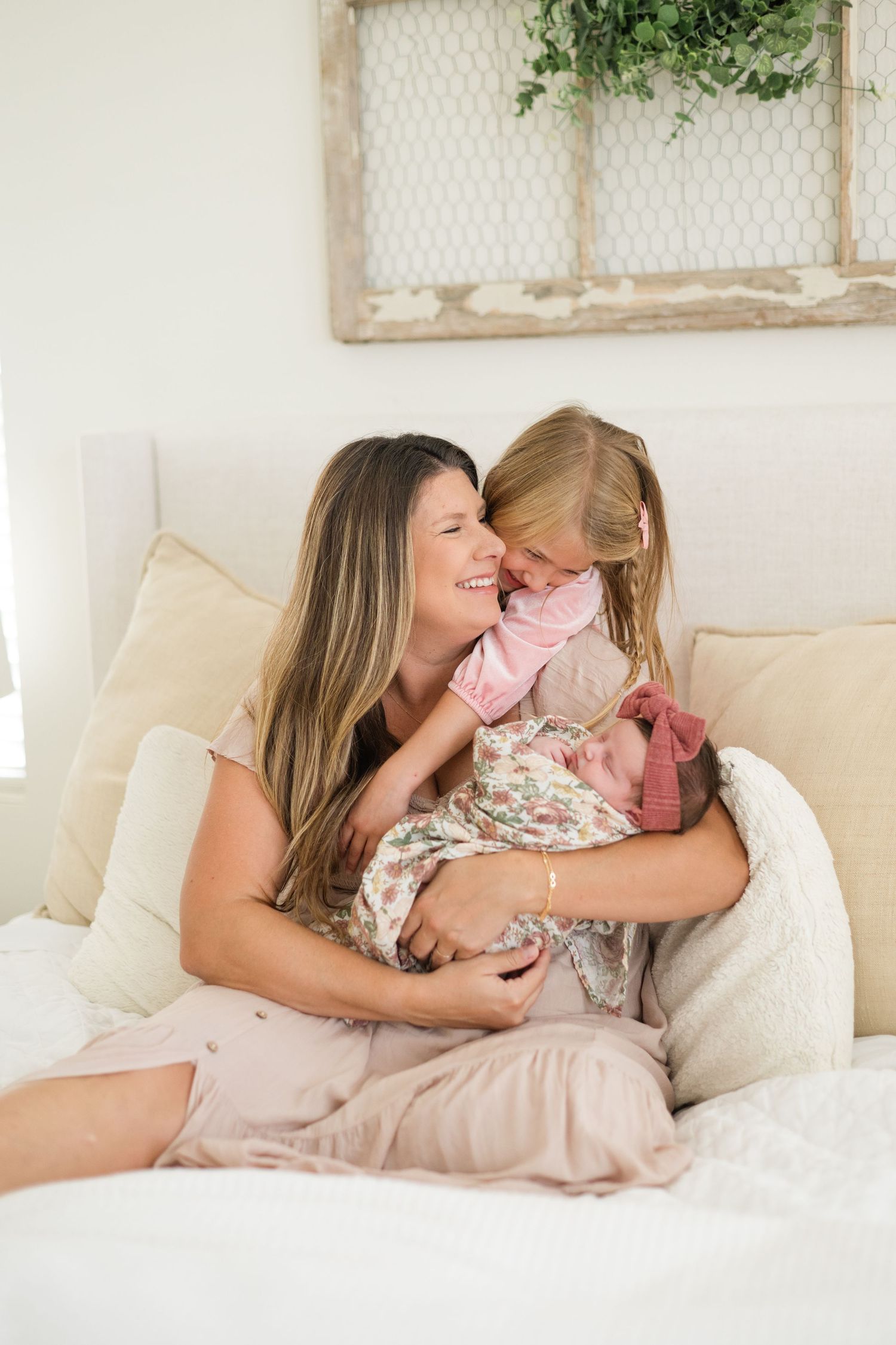 Mother smiling while holding newborn baby as young blonde daughter leans in to kiss sibling on white bed.