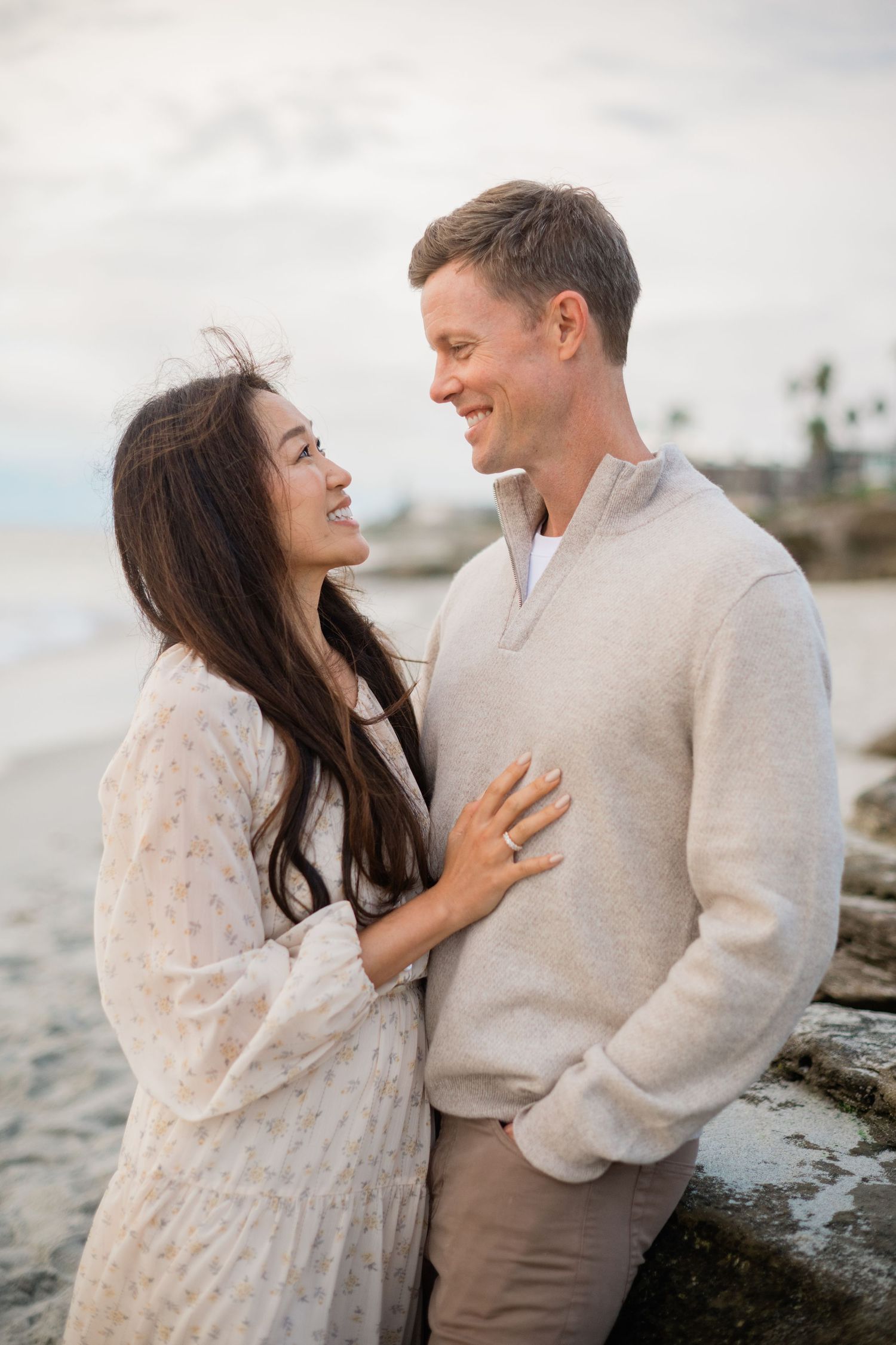 Smiling couple shares a joyful moment together on a beach, woman in floral dress and man in beige sweater.