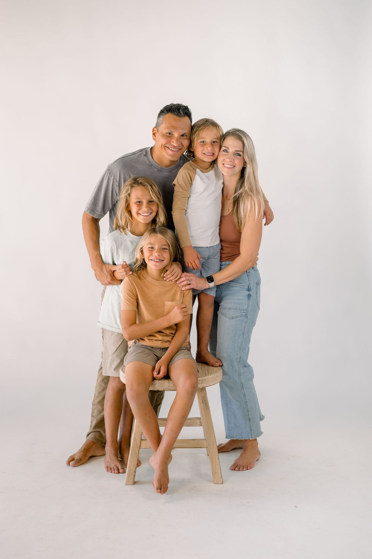 A happy family of five poses together in a bright white studio, smiling warmly in casual neutral-toned outfits.