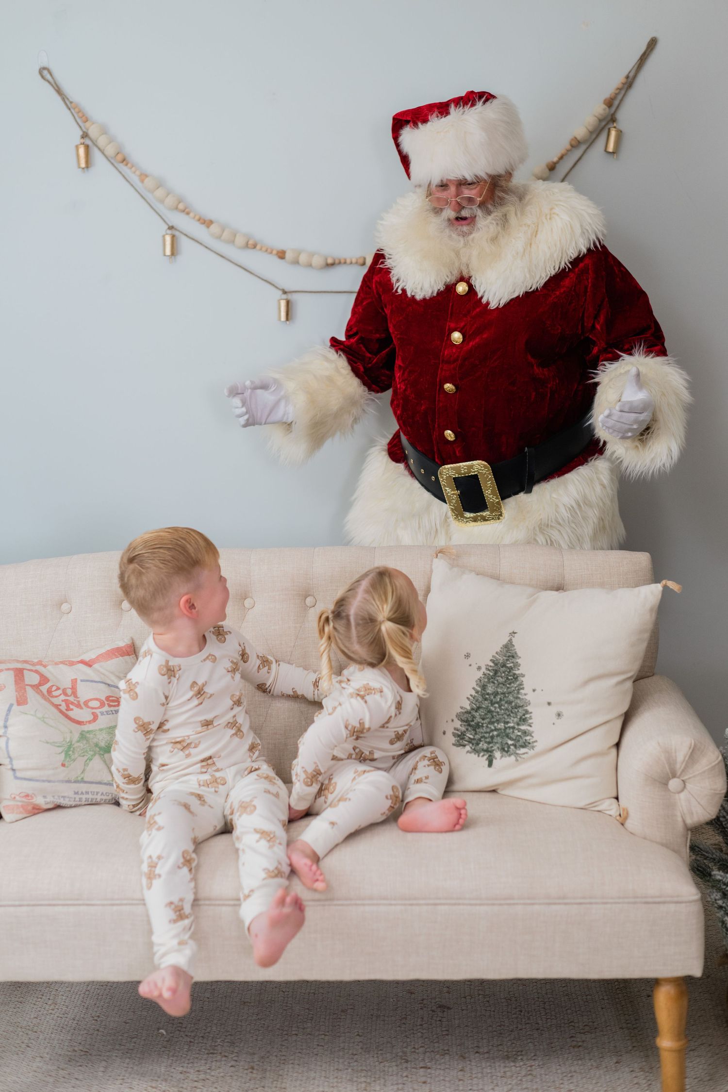 Two toddlers in pajamas sit on sofa as Santa Claus gestures behind them at Christmas.