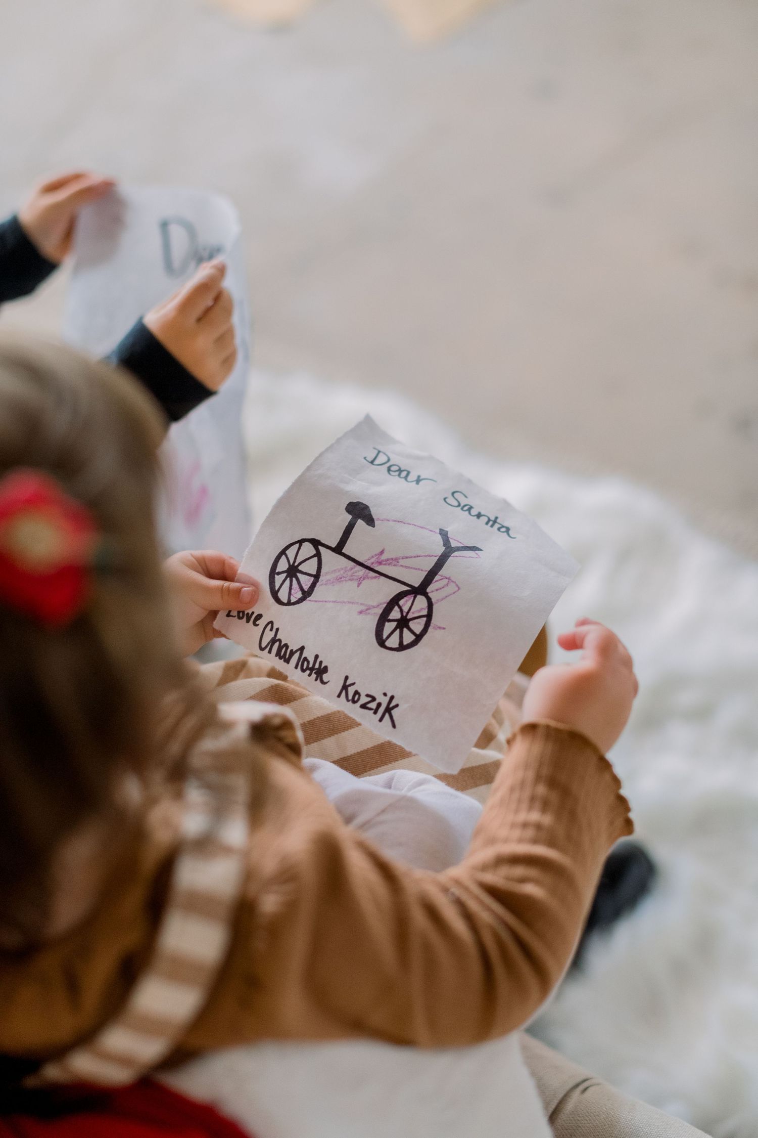 A child holds a hand-drawn letter to Santa Claus featuring a bicycle, signed Love Charlotte Kozik.