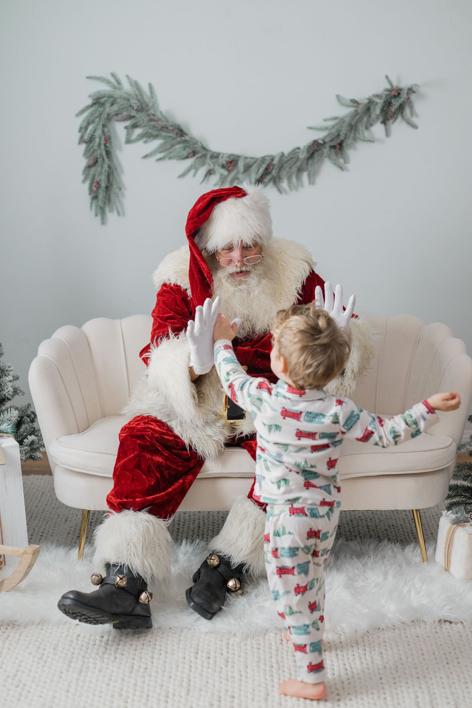 A toddler in festive pajamas reaches out to Santa Claus sitting on a cream sofa in a Christmas decorated room.