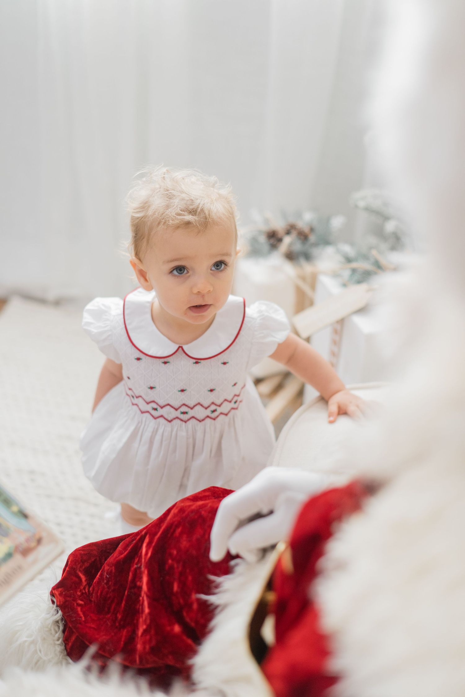 Blonde toddler girl in white smocked dress with red trim looks up at Santa Claus during Christmas photo session.