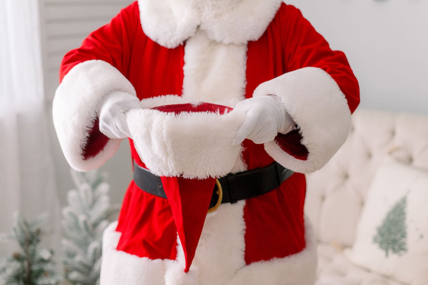 Santa Claus in red suit and white gloves holding his hat indoors with Christmas decorations in background.