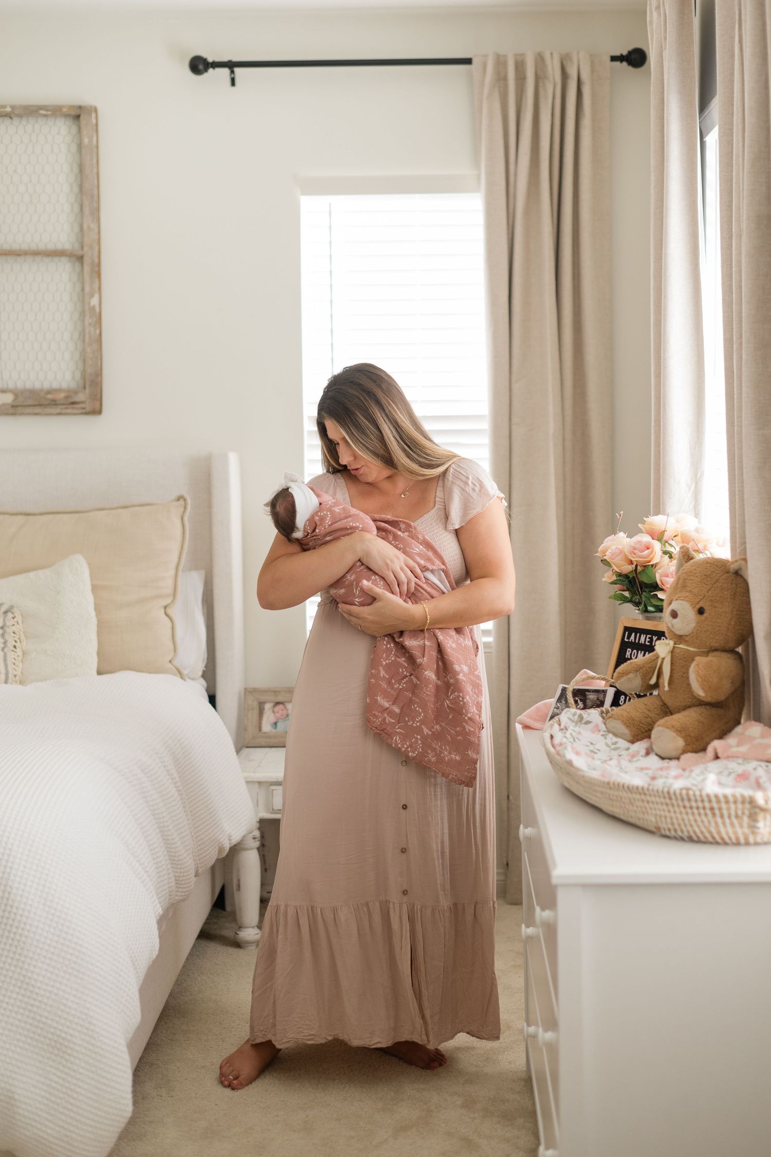 A woman in a maxi dress holds her newborn baby in a pink swaddle blanket in a softly decorated bedroom.