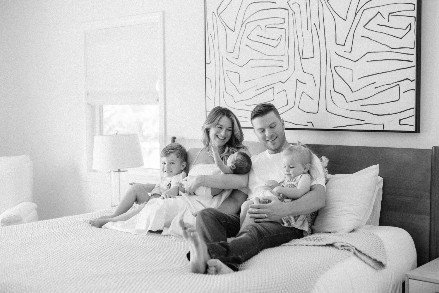 Black and white family portrait of parents with newborn and toddler sitting together on a bed.