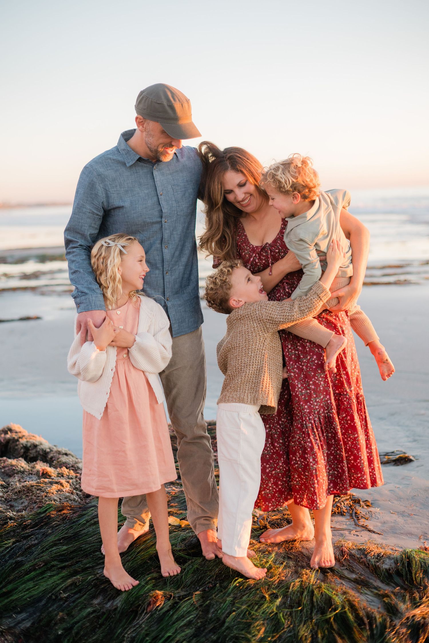 Family of five sharing a warm embrace on a rocky beach at golden hour sunset.