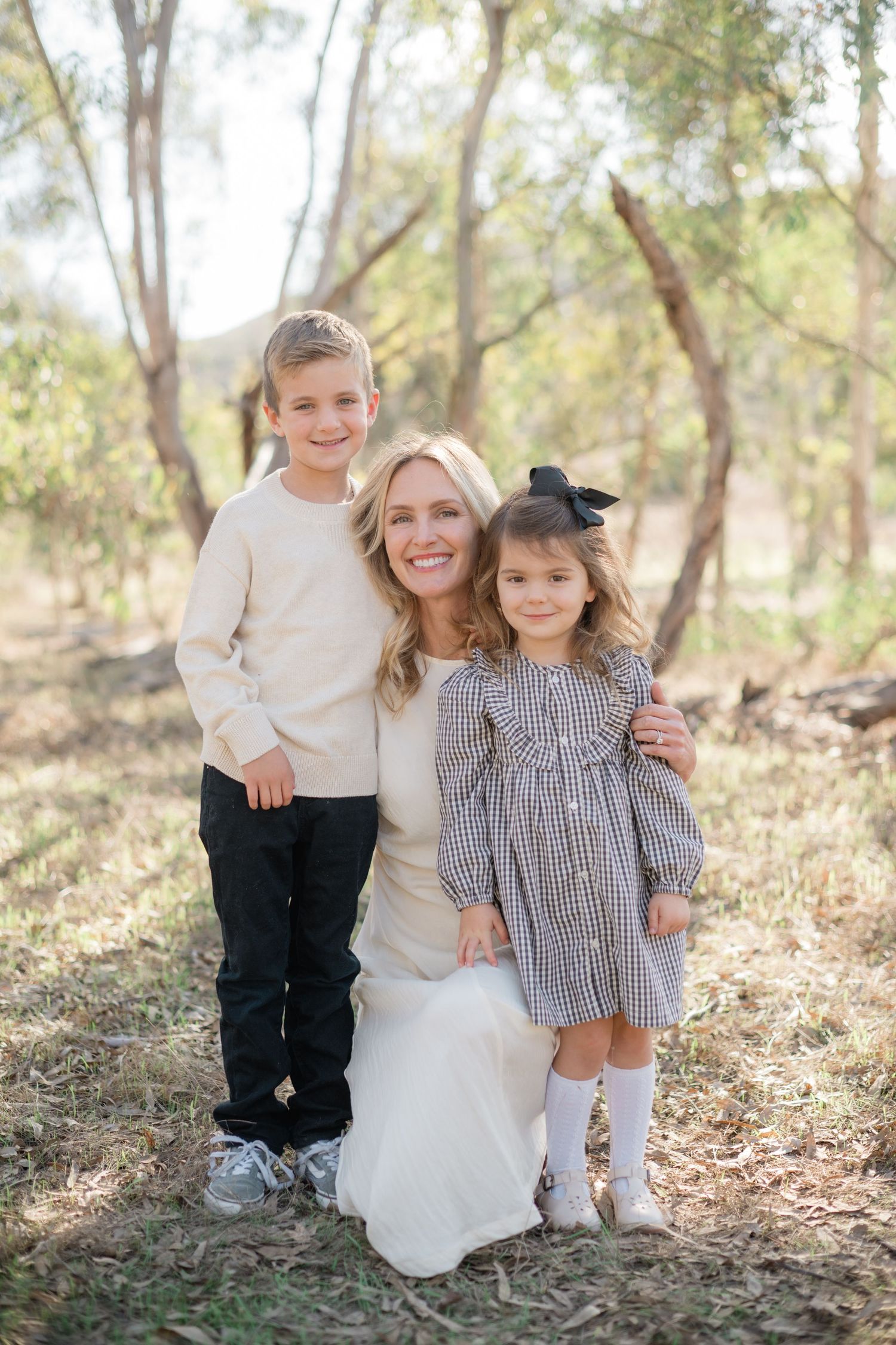 A smiling blonde mother kneels outdoors with her two young children in a sunlit woodland setting.