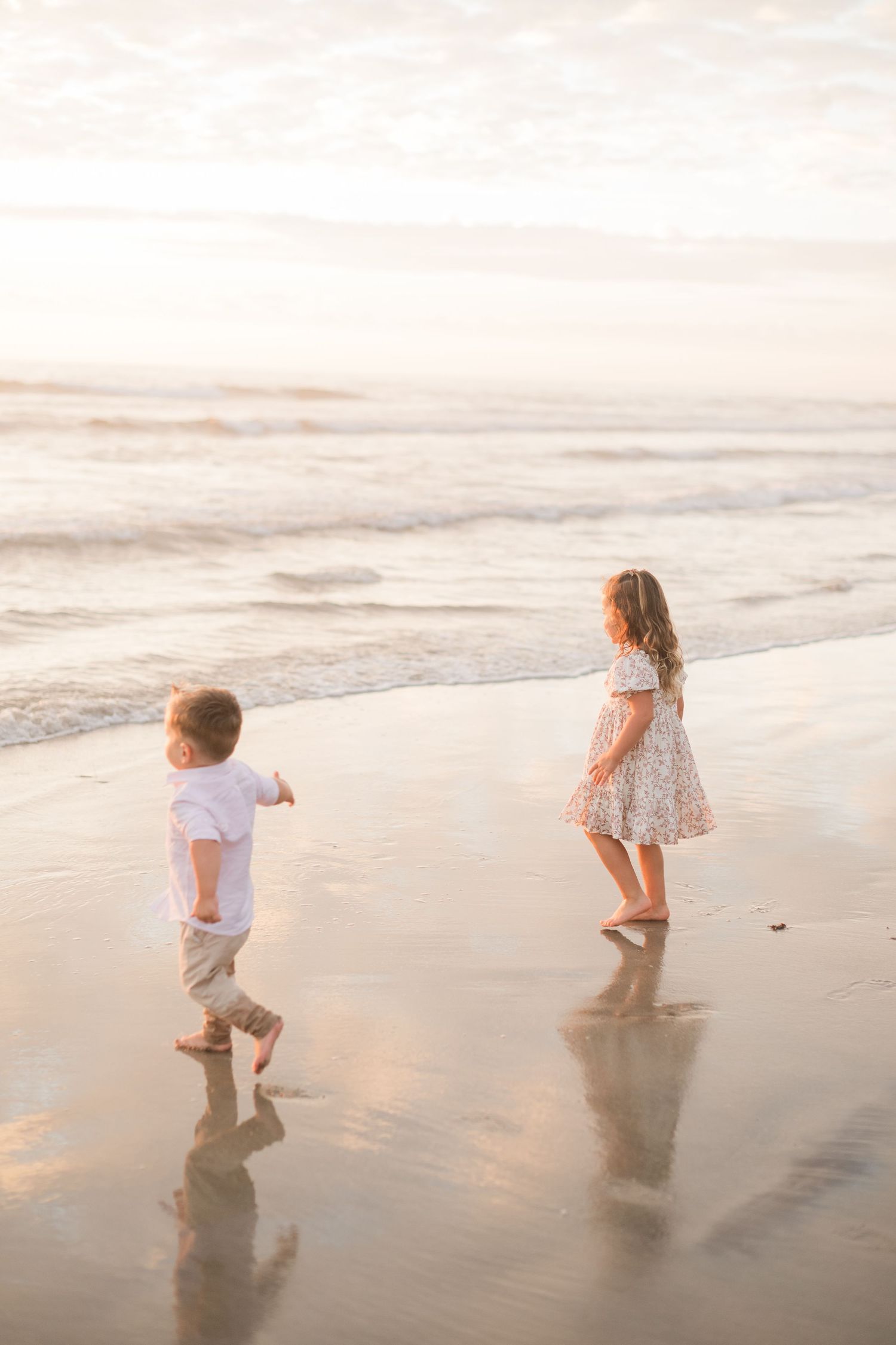 Two young children playing barefoot on a sandy beach at sunset, with gentle ocean waves in the background.