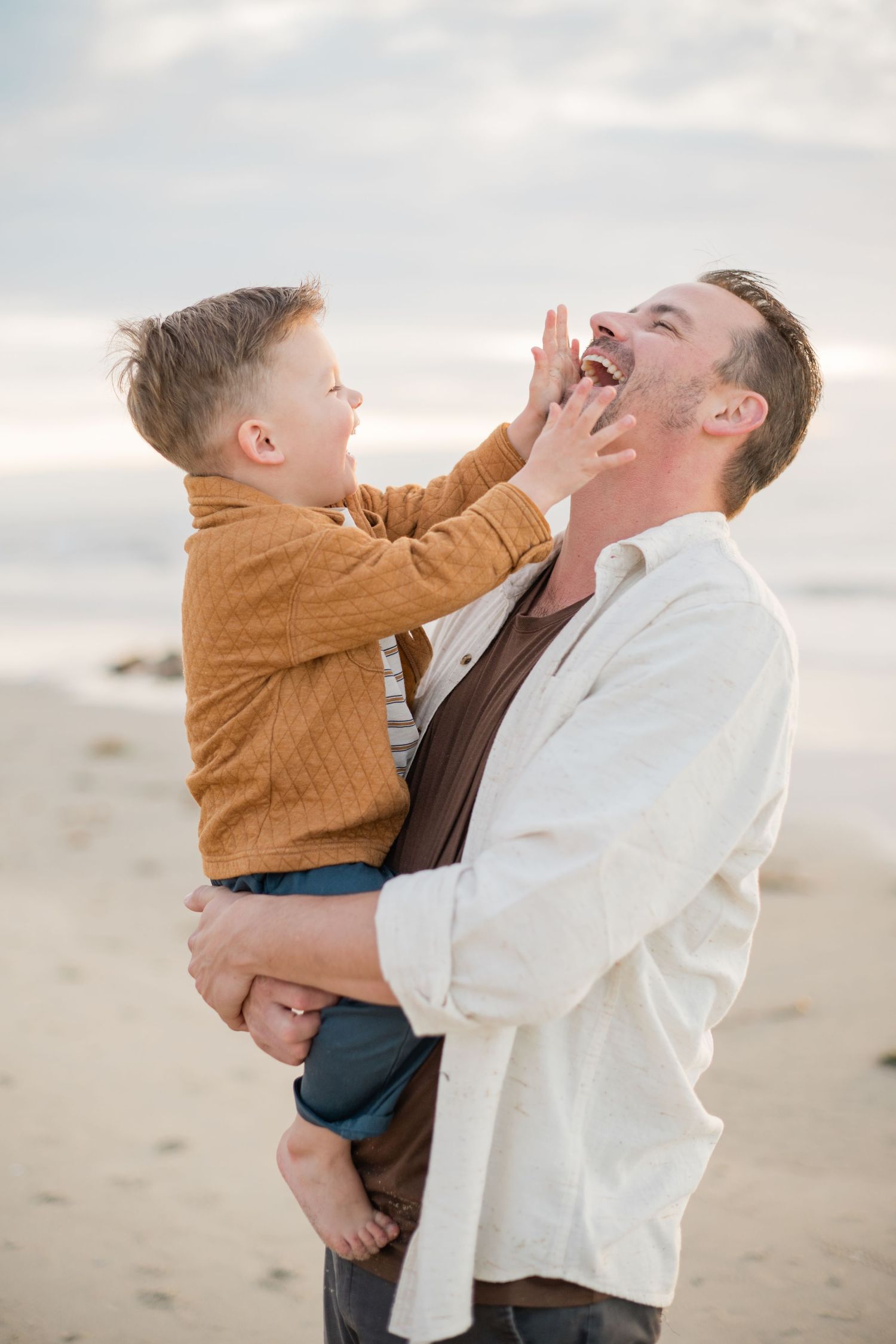 Laughing father holds young boy in brown jacket on beach as child playfully touches dad's face.