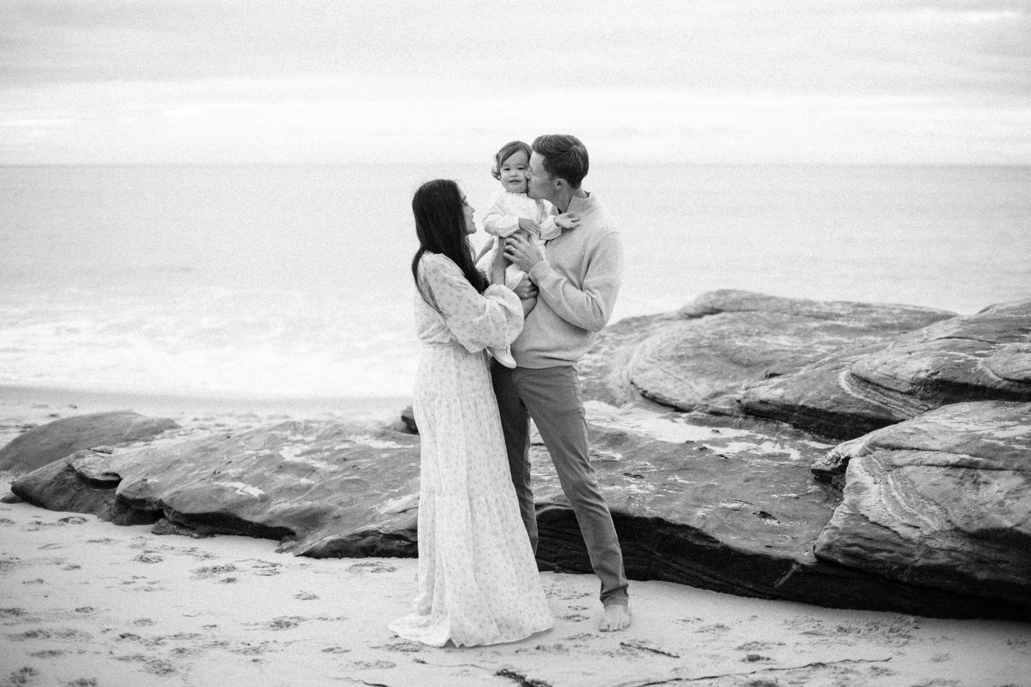 Black and white beach photo of a couple holding their baby near rocky shoreline with ocean waves in the background.